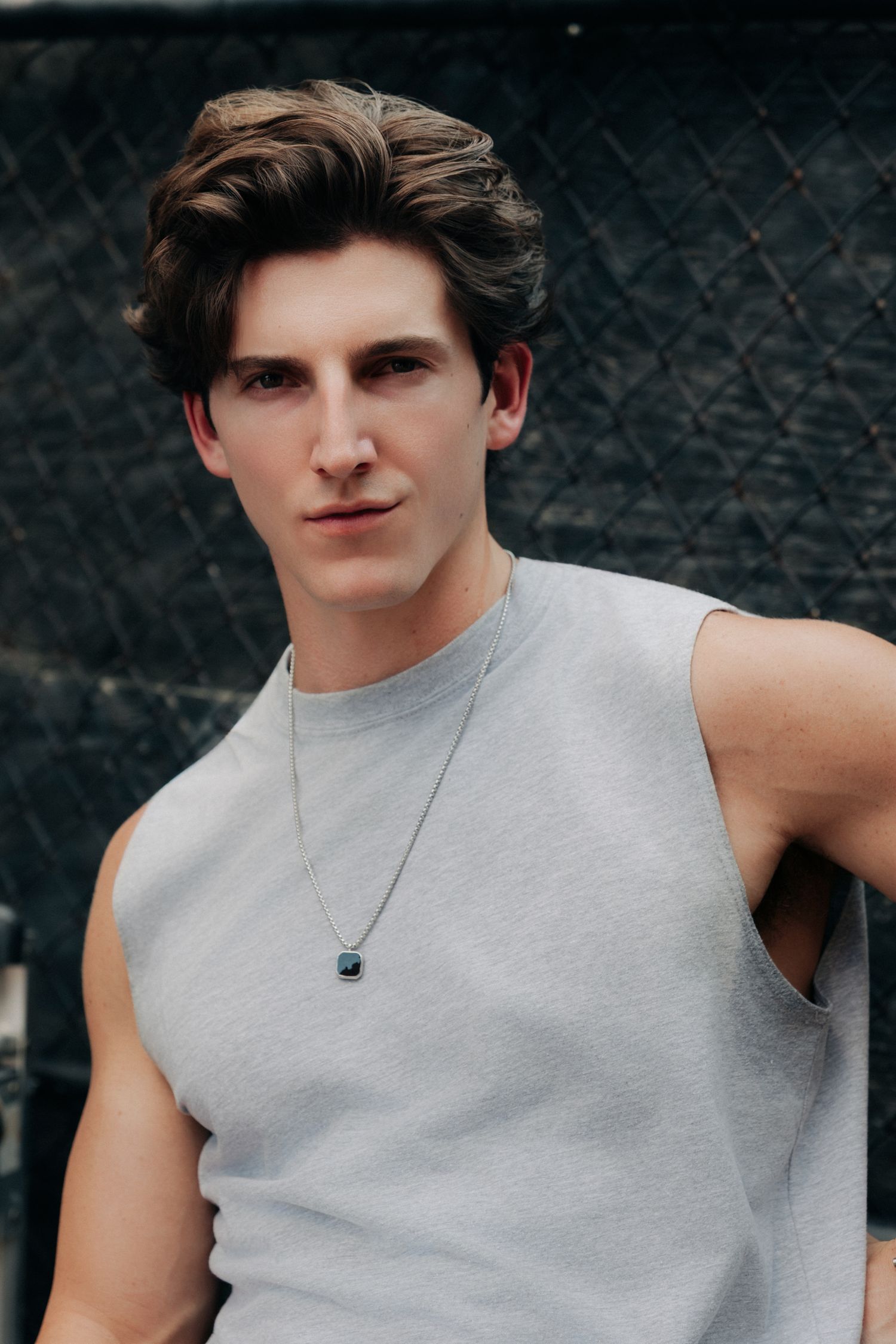 Young man in gray sleeveless shirt wearing a pendant necklace, posing confidently against a dark chain-link fence.