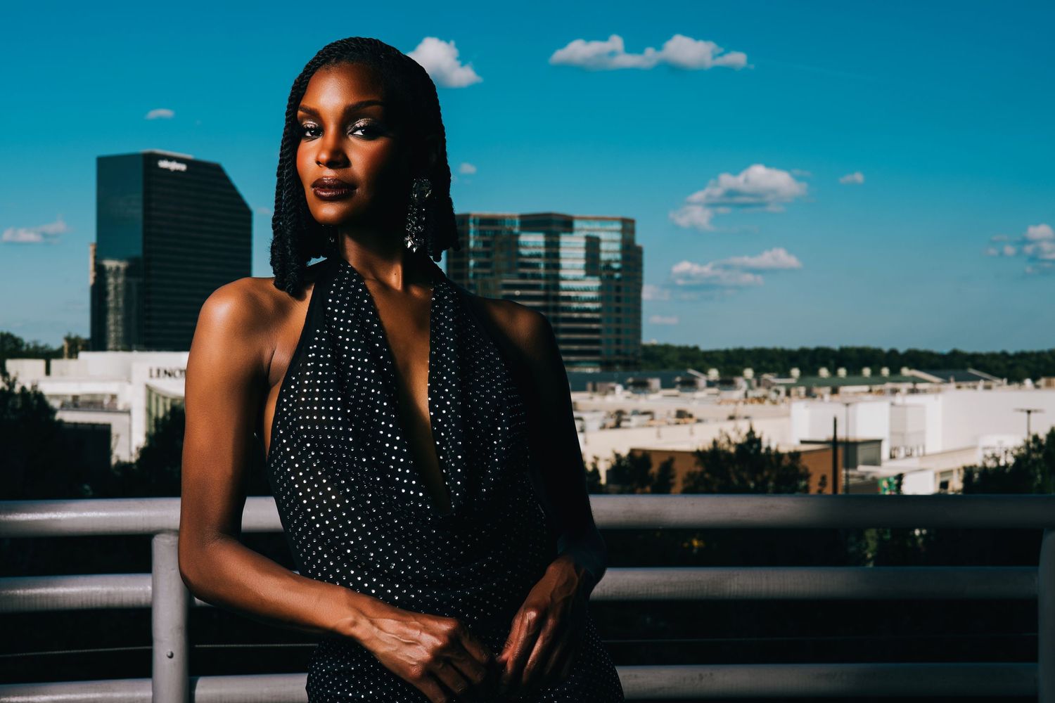 A stylish woman in a black polka dot halter dress poses on a rooftop with a city skyline behind her.