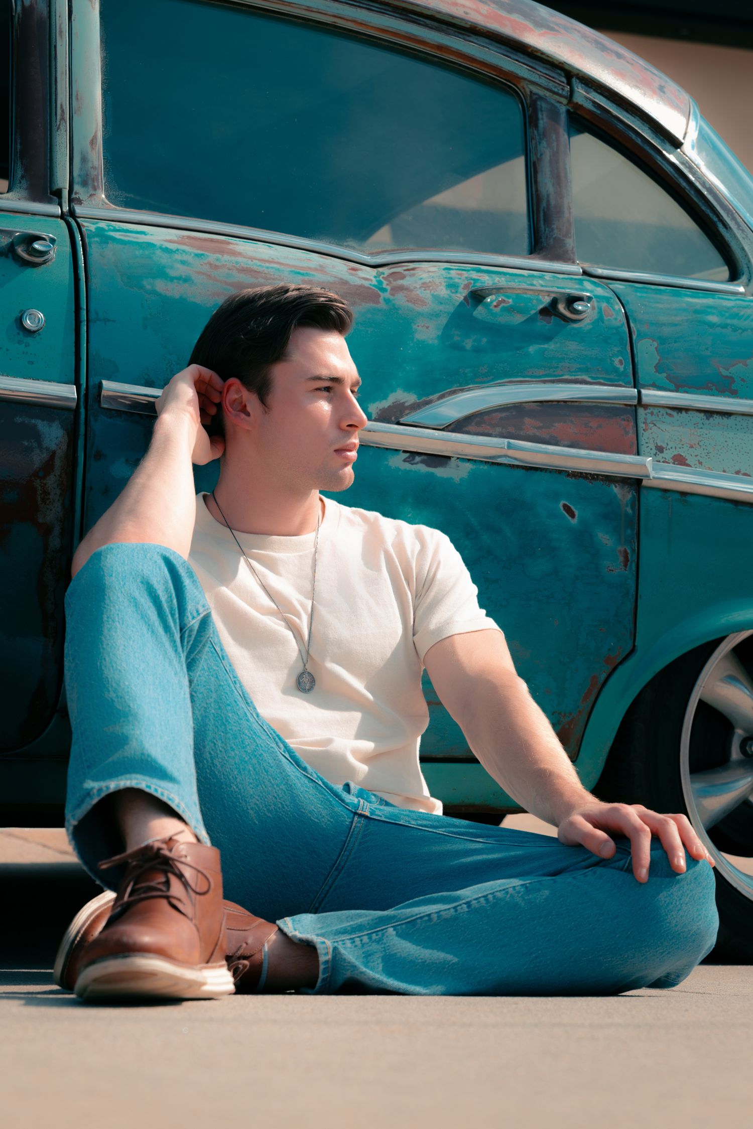 Young man in white tee and jeans sits cross-legged against a vintage turquoise car, gazing thoughtfully to the side.