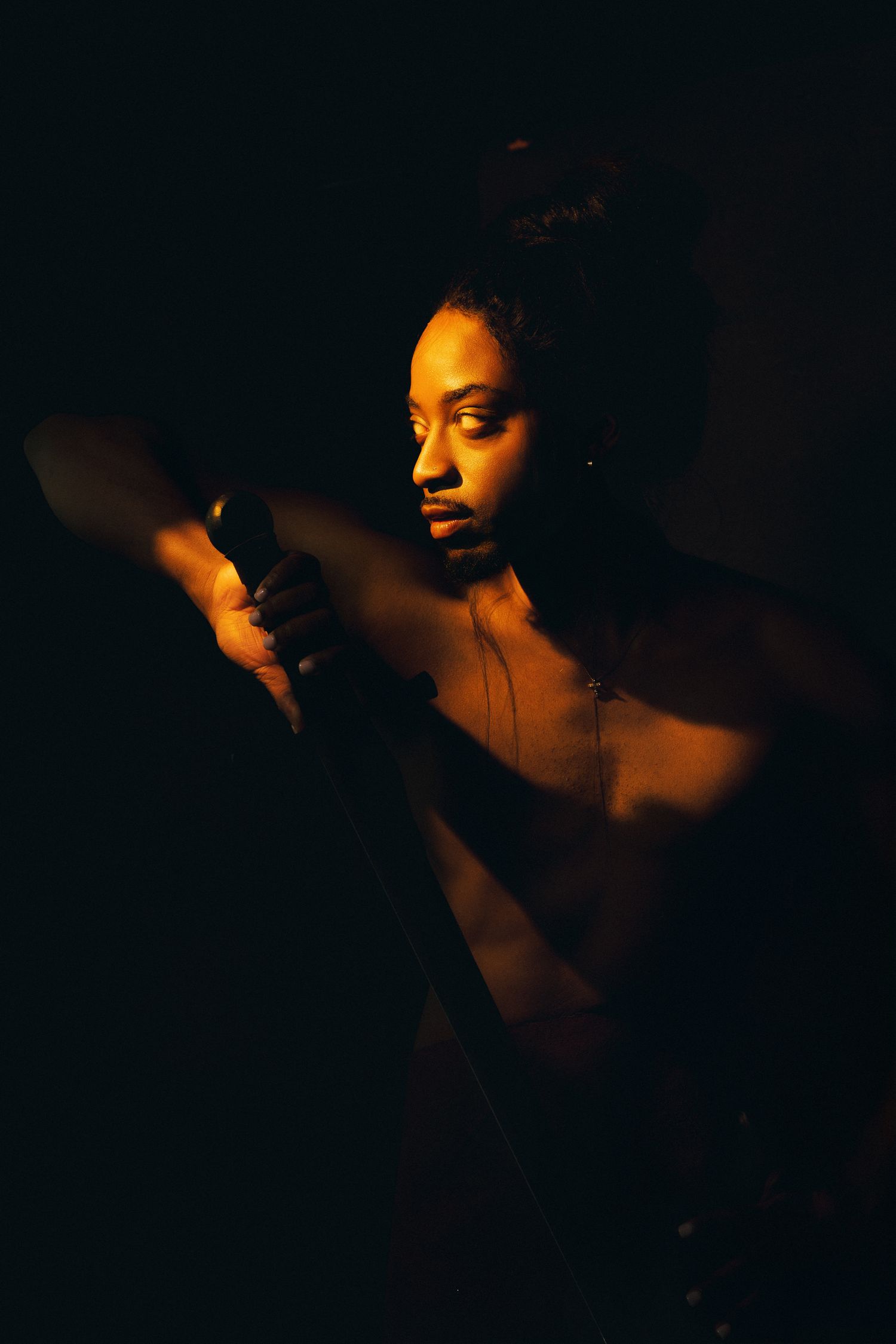 A person illuminated by warm golden light against a dark background, wearing a necklace, in a dramatic artistic portrait.