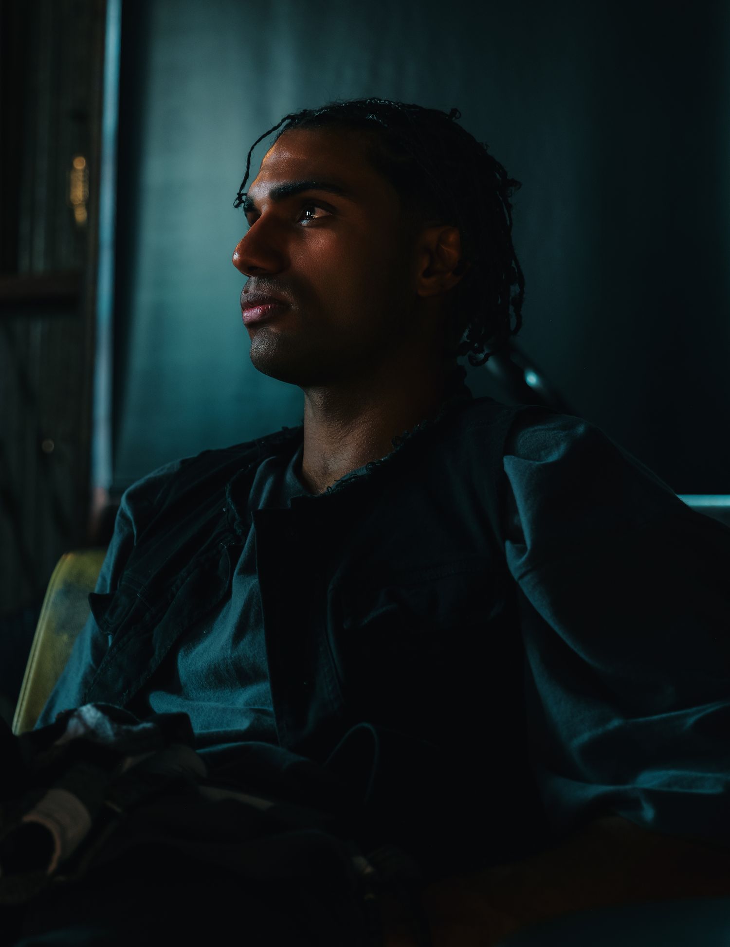 Young man with braided hair sitting in dim blue-toned lighting, gazing thoughtfully upward in a dark room.