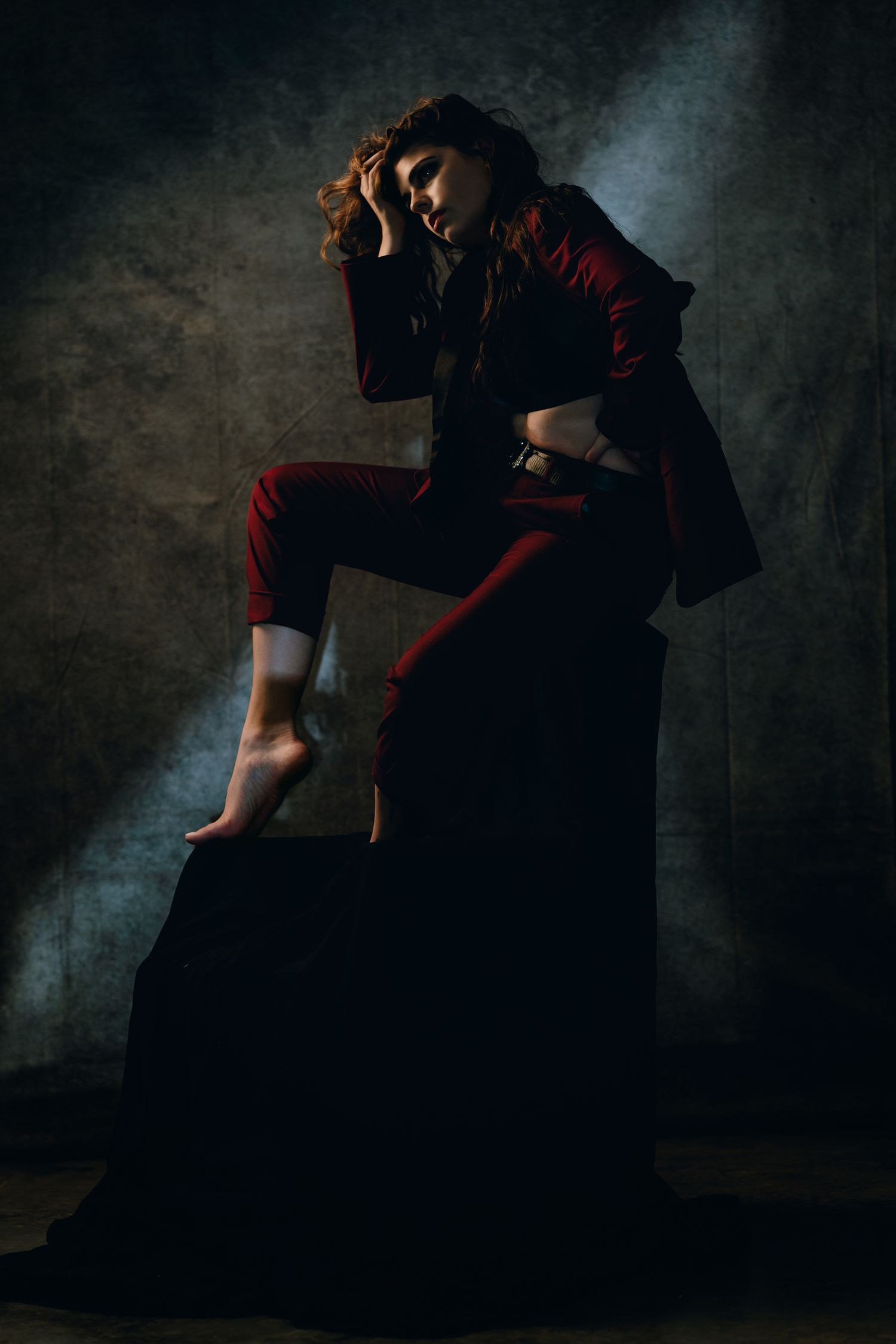 A dramatic fashion portrait of a woman in red pants and jacket posed on a dark cube against a moody gray background.