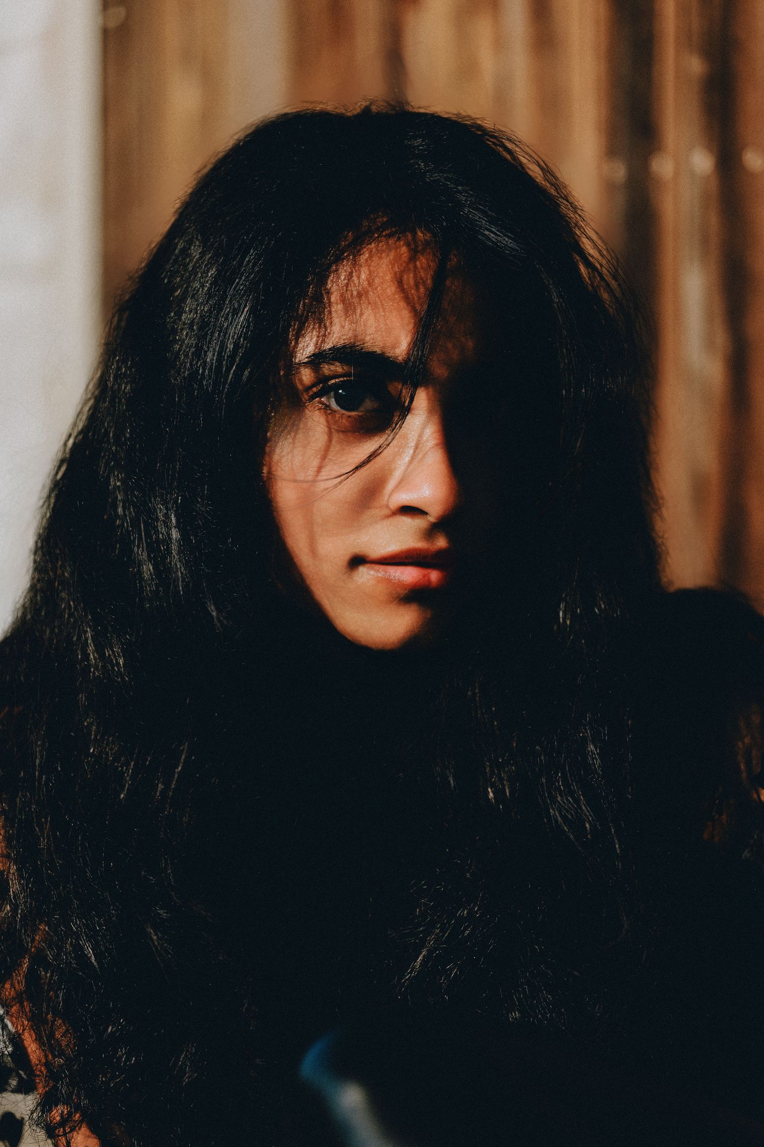 A young person with long dark wavy hair poses in dramatic moody lighting against a wooden background.