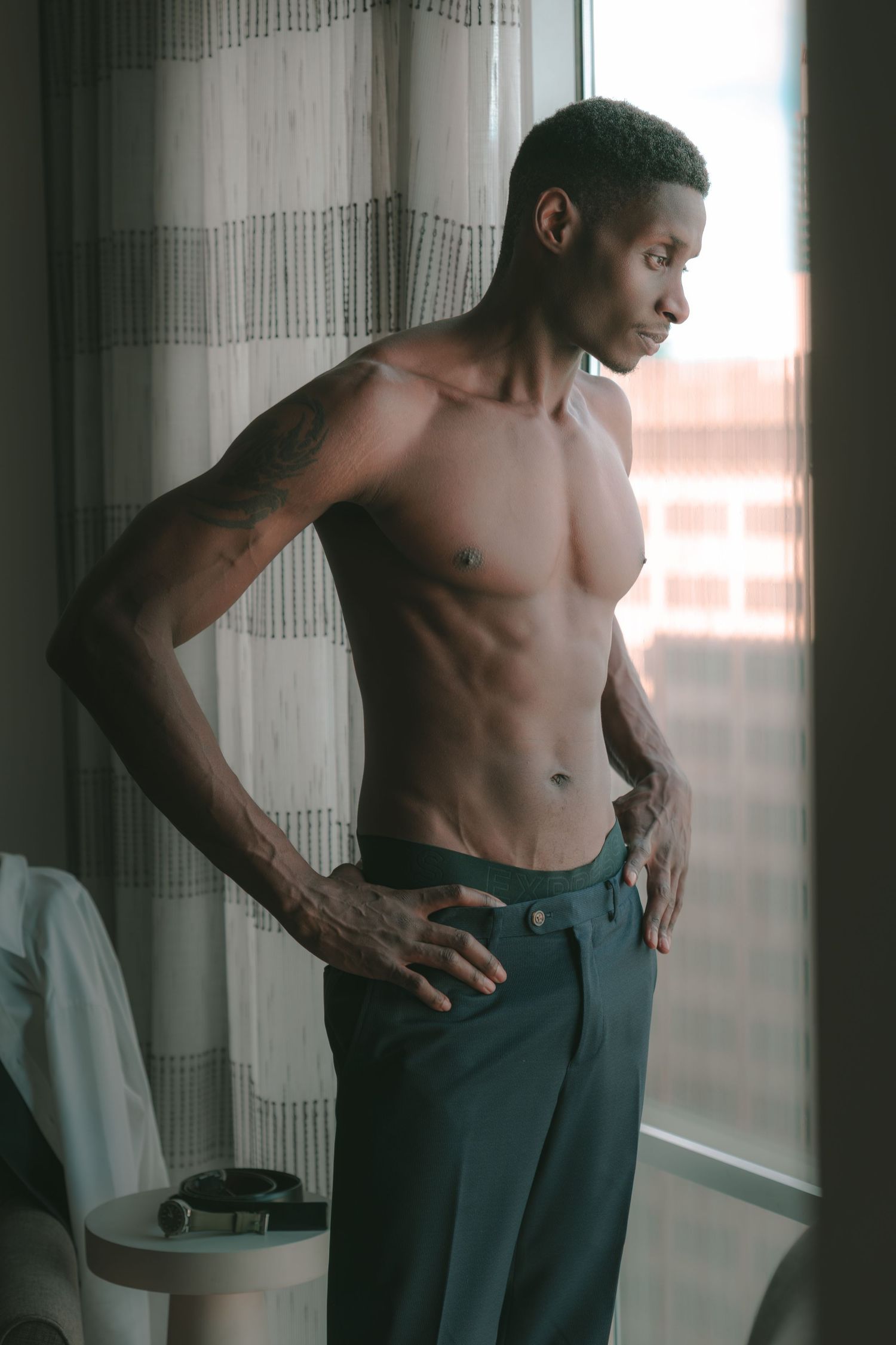 A shirtless muscular Black man with tattoos stands by a window in gray trousers, gazing outside in natural light.