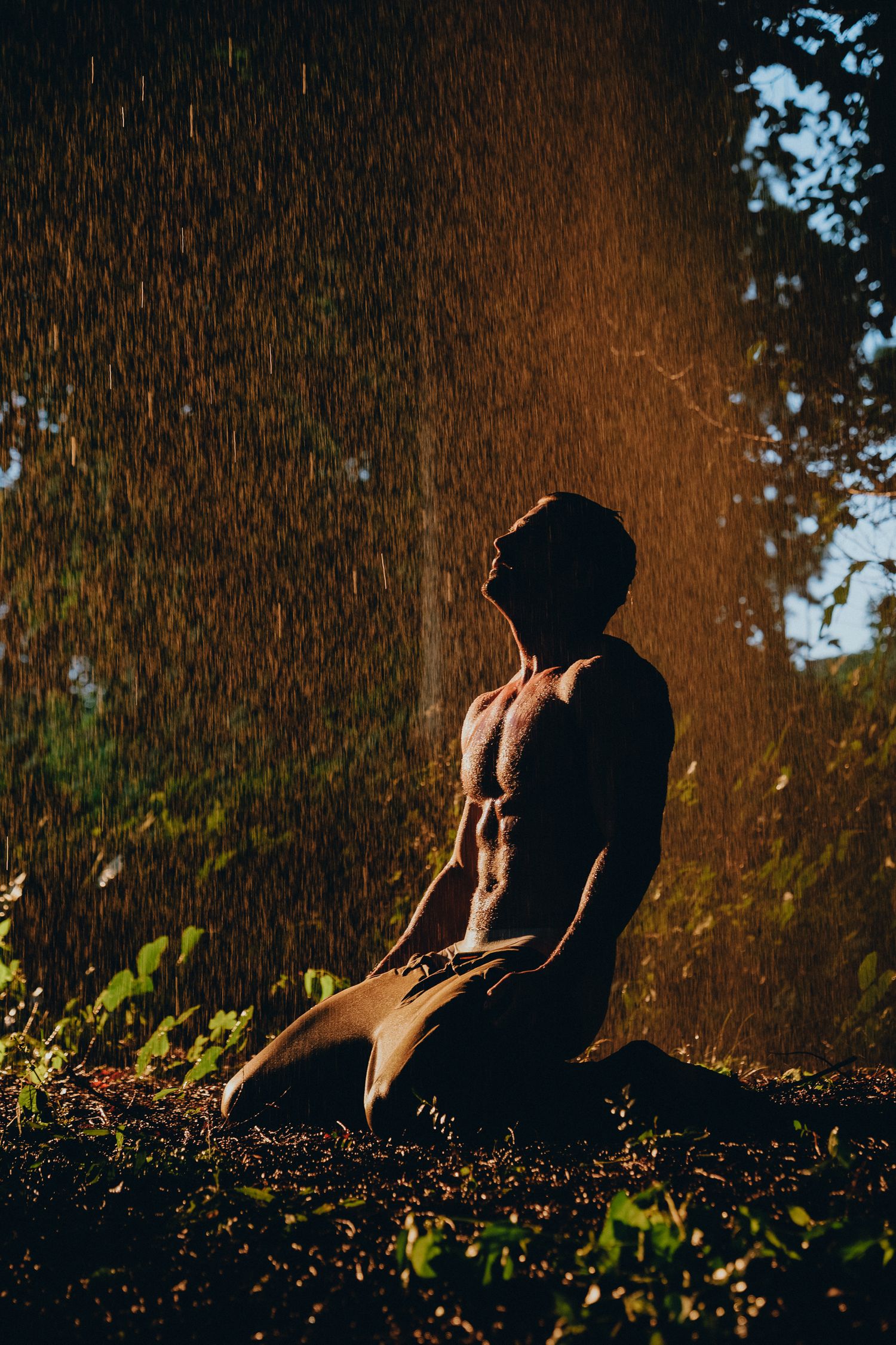 Muscular man kneeling shirtless in rain outdoors, backlit by golden sunlight with lush green trees in background.