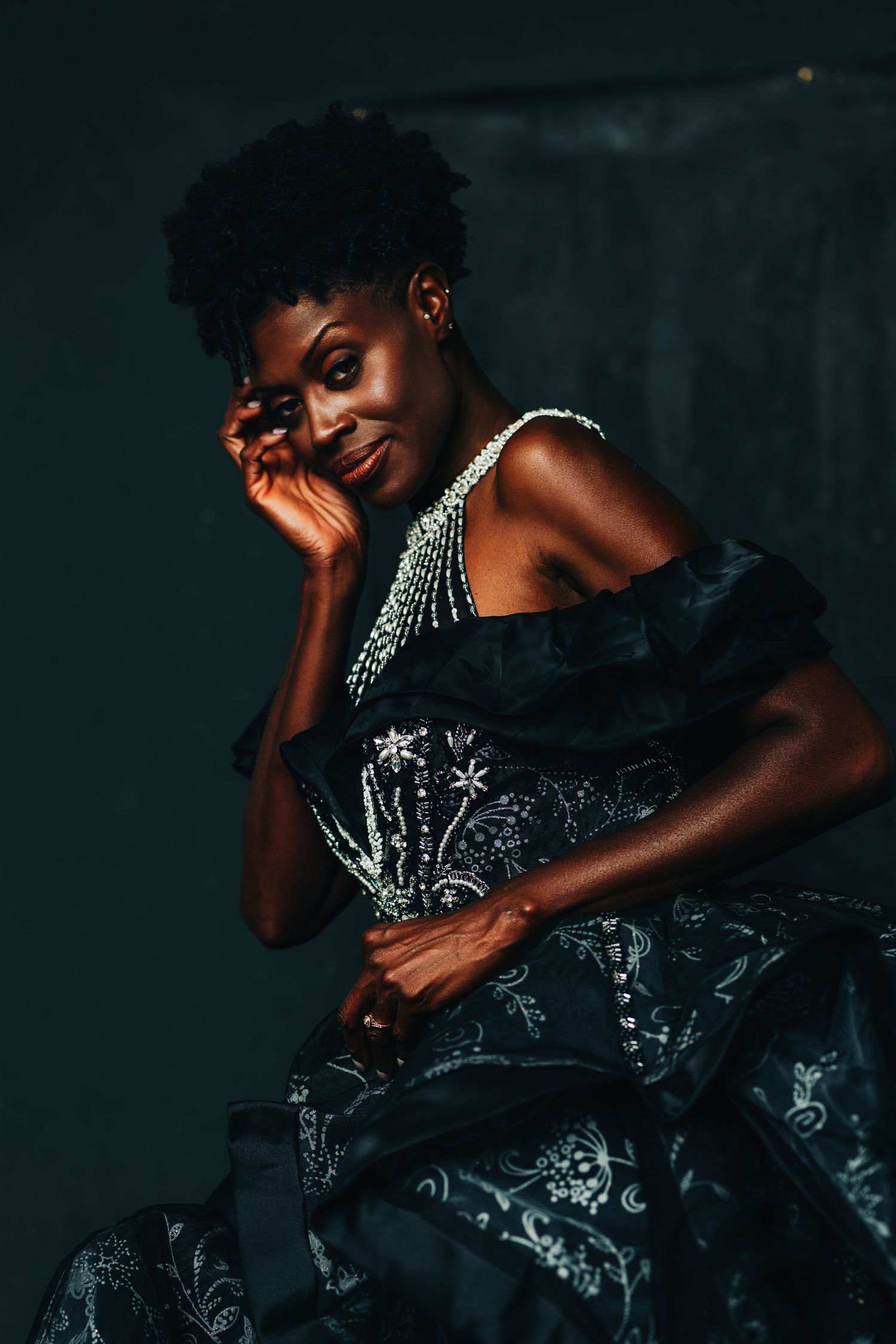 Elegant Black woman in embroidered off-shoulder gown with pearl necklace poses gracefully against dark background.