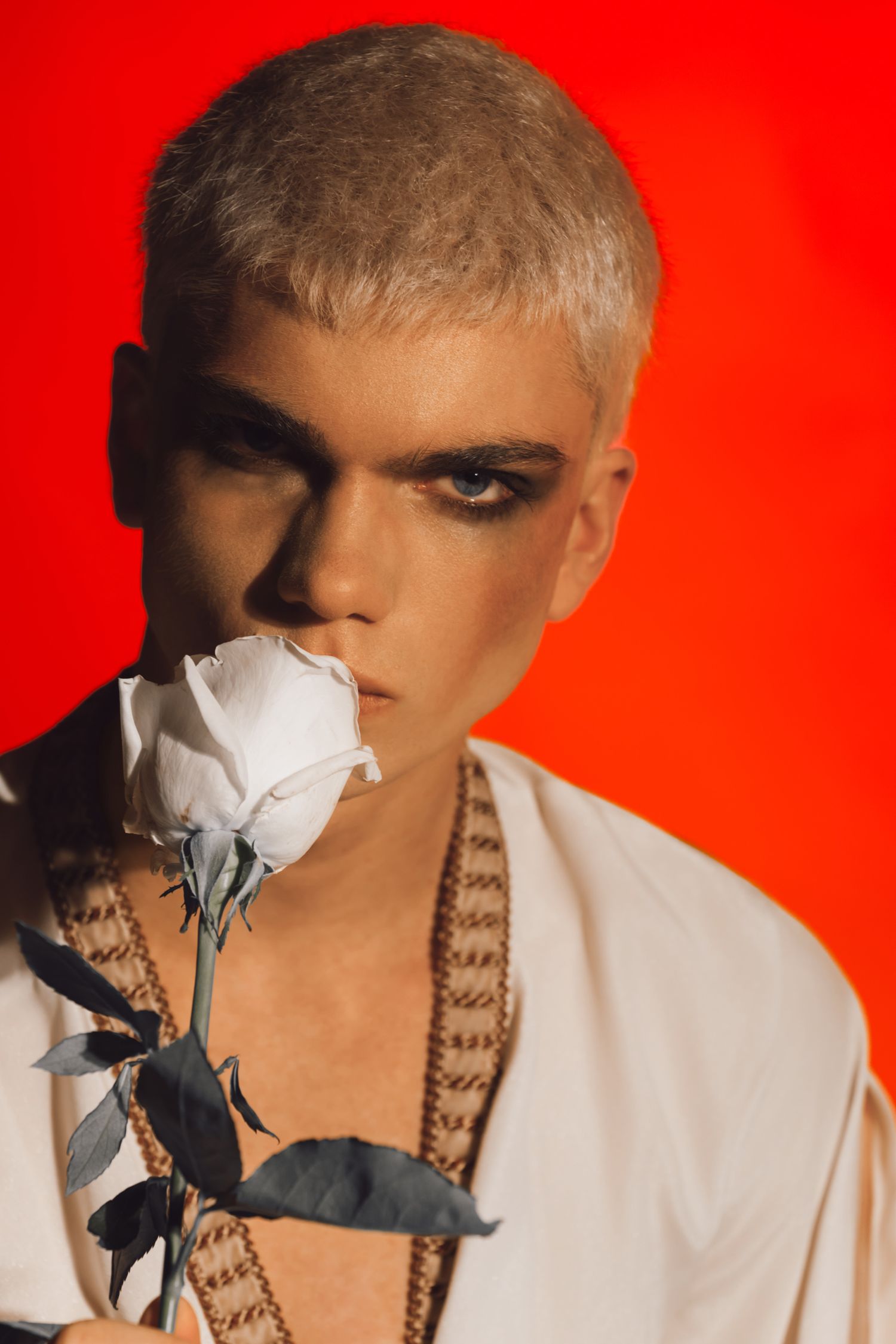 Young man with blonde hair and dark eye makeup holds a white rose against a bold red background.