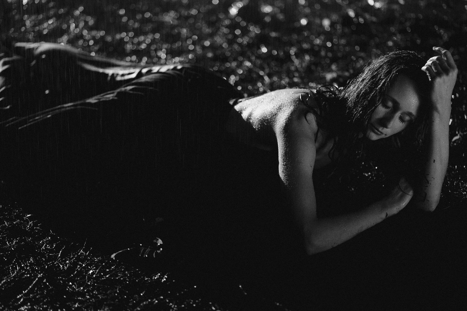 Black and white photo of a woman in a dark dress lying on the ground in a dramatic, moody outdoor setting.