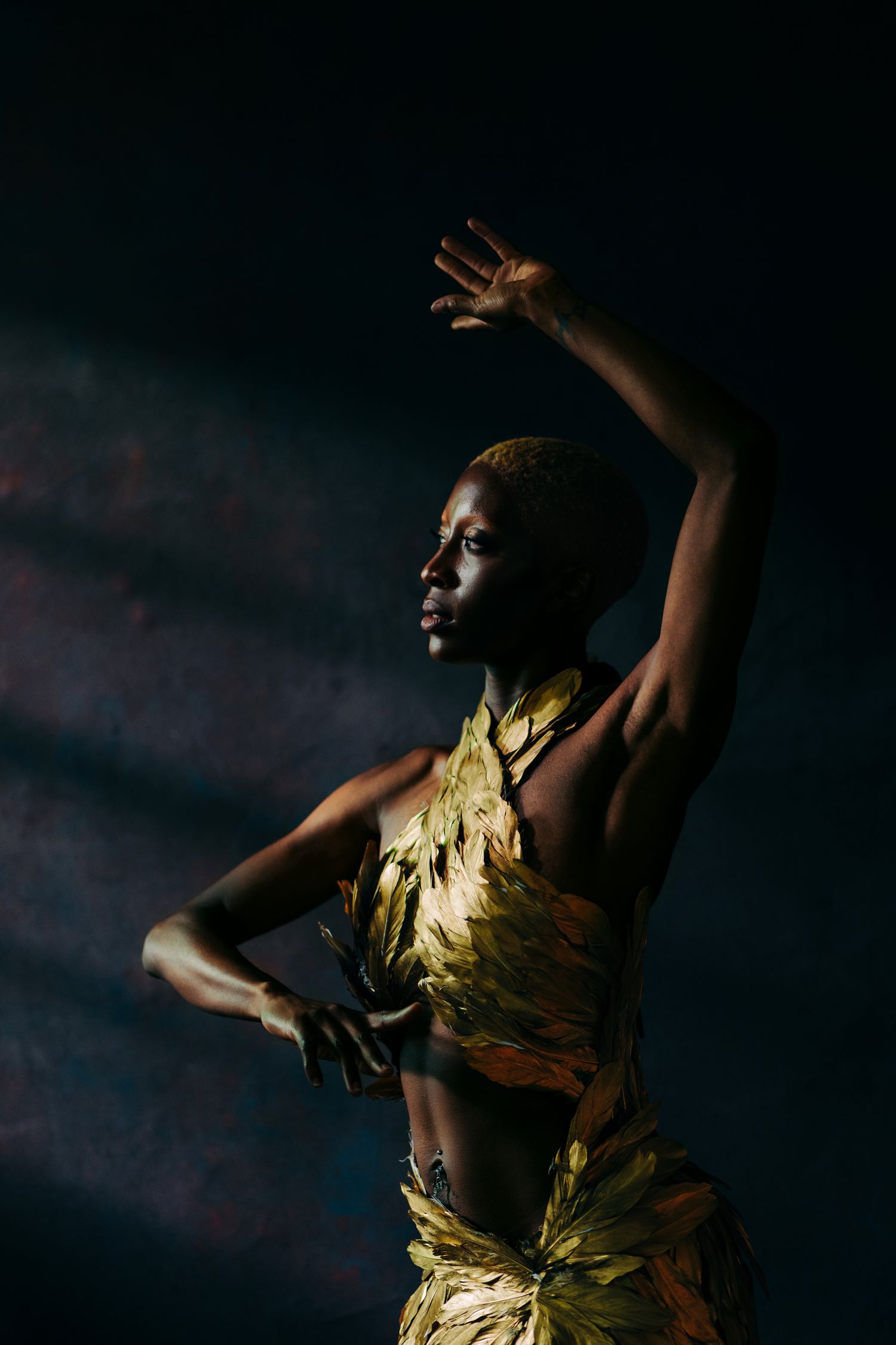A striking model in a golden feathered outfit poses dramatically against a dark background with dramatic lighting.