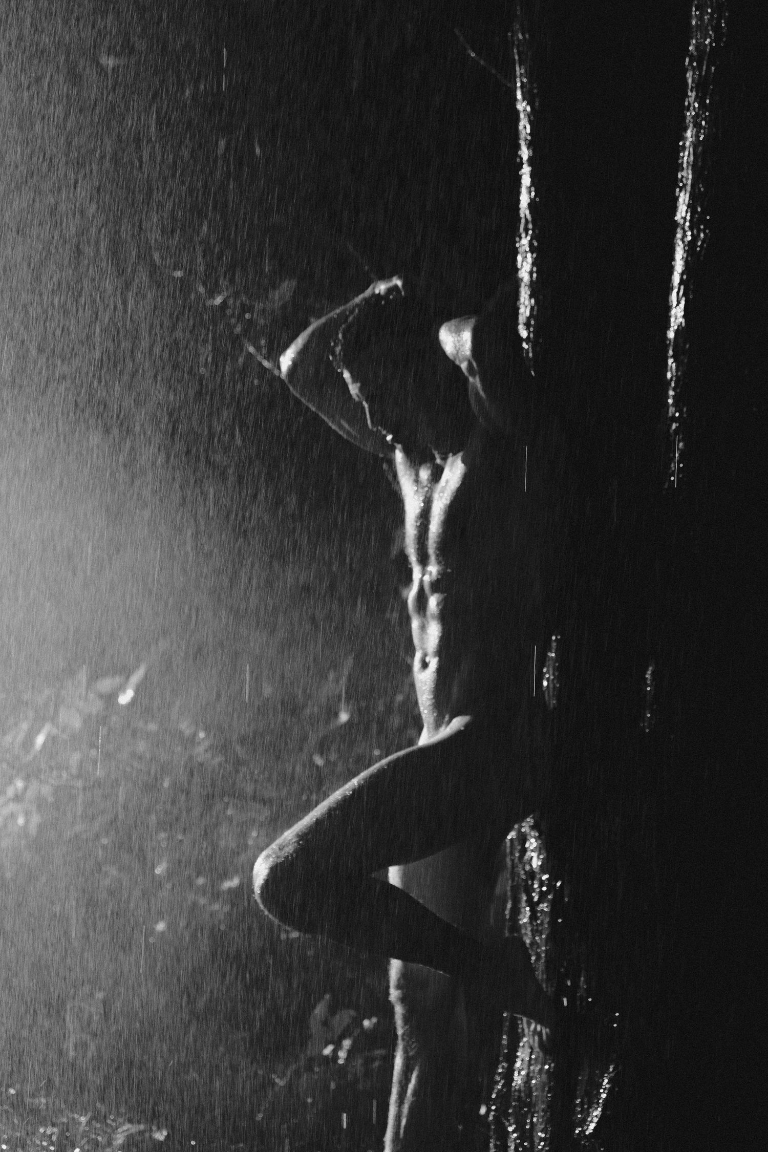 Black and white photo of a muscular man standing in the rain against a dark rocky wall.