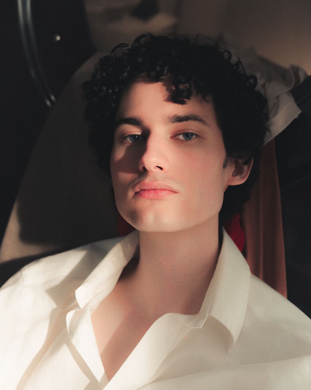 Young man with dark curly hair wearing a white button-up shirt, posed in soft dramatic lighting.