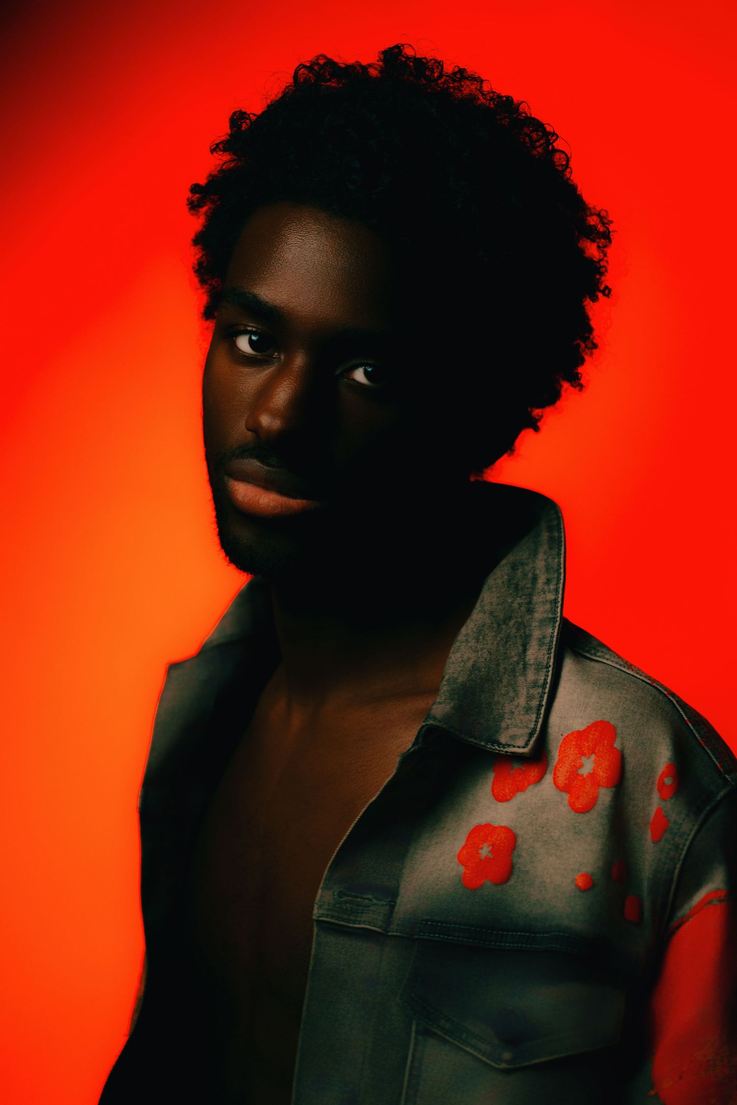 Young Black man in floral denim jacket poses against vibrant red background in artistic fashion portrait.