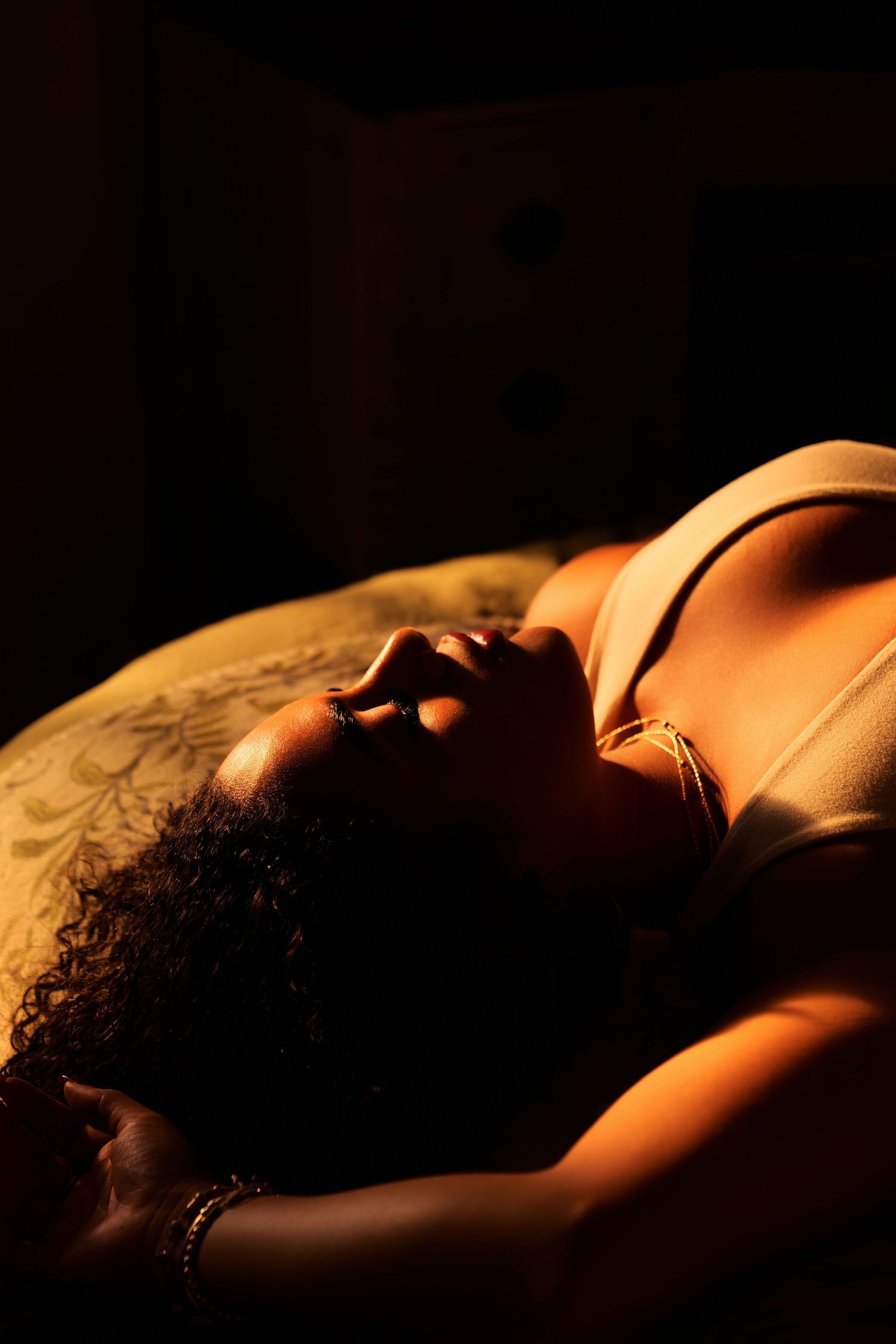 A woman with curly hair reclines in warm golden light wearing a bralette, captured in dramatic chiaroscuro lighting.