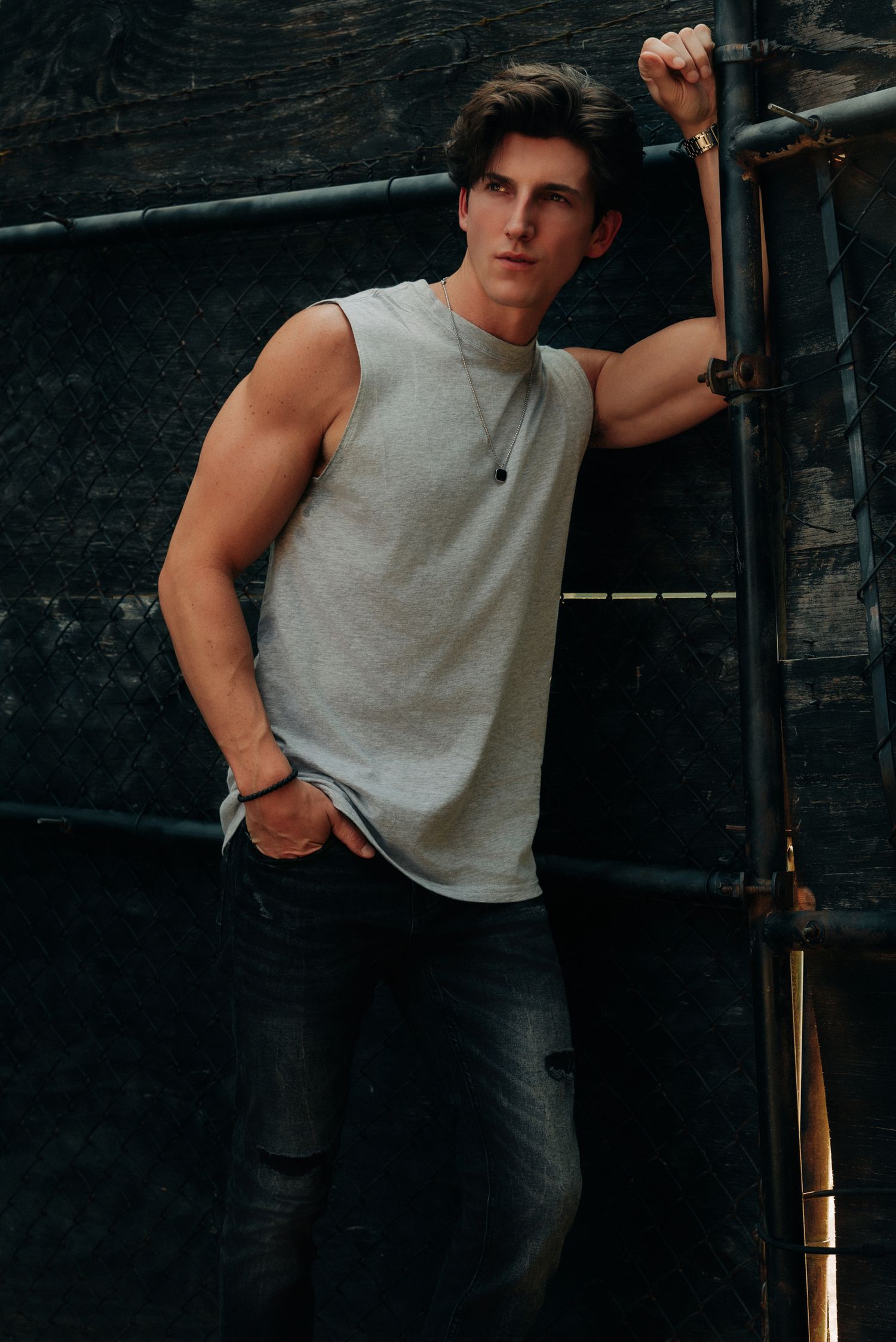 Athletic young man in gray sleeveless shirt and ripped jeans leaning against metal scaffolding outdoors.