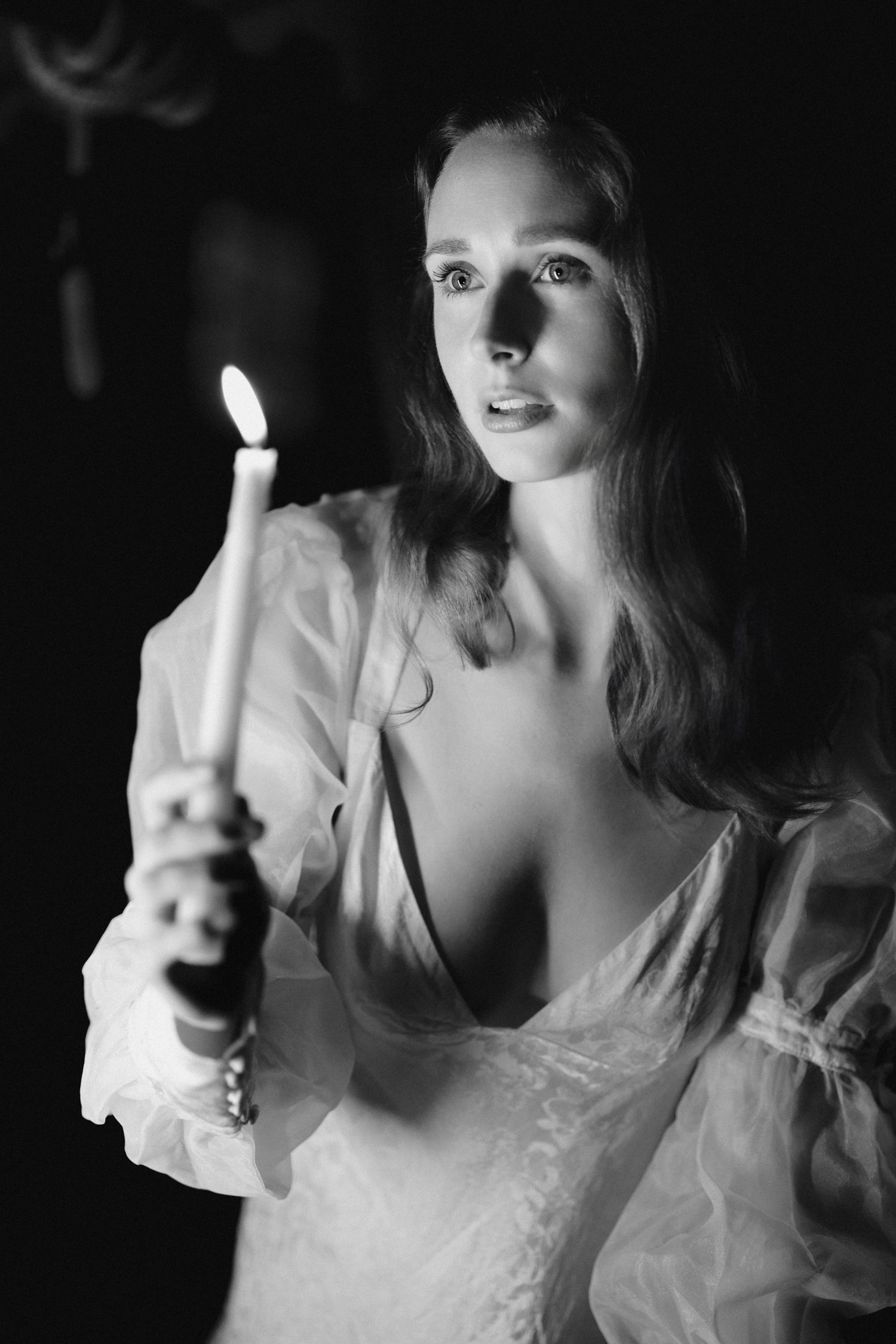 Black and white portrait of a woman holding a lit candle in dim light, wearing a flowing white dress.