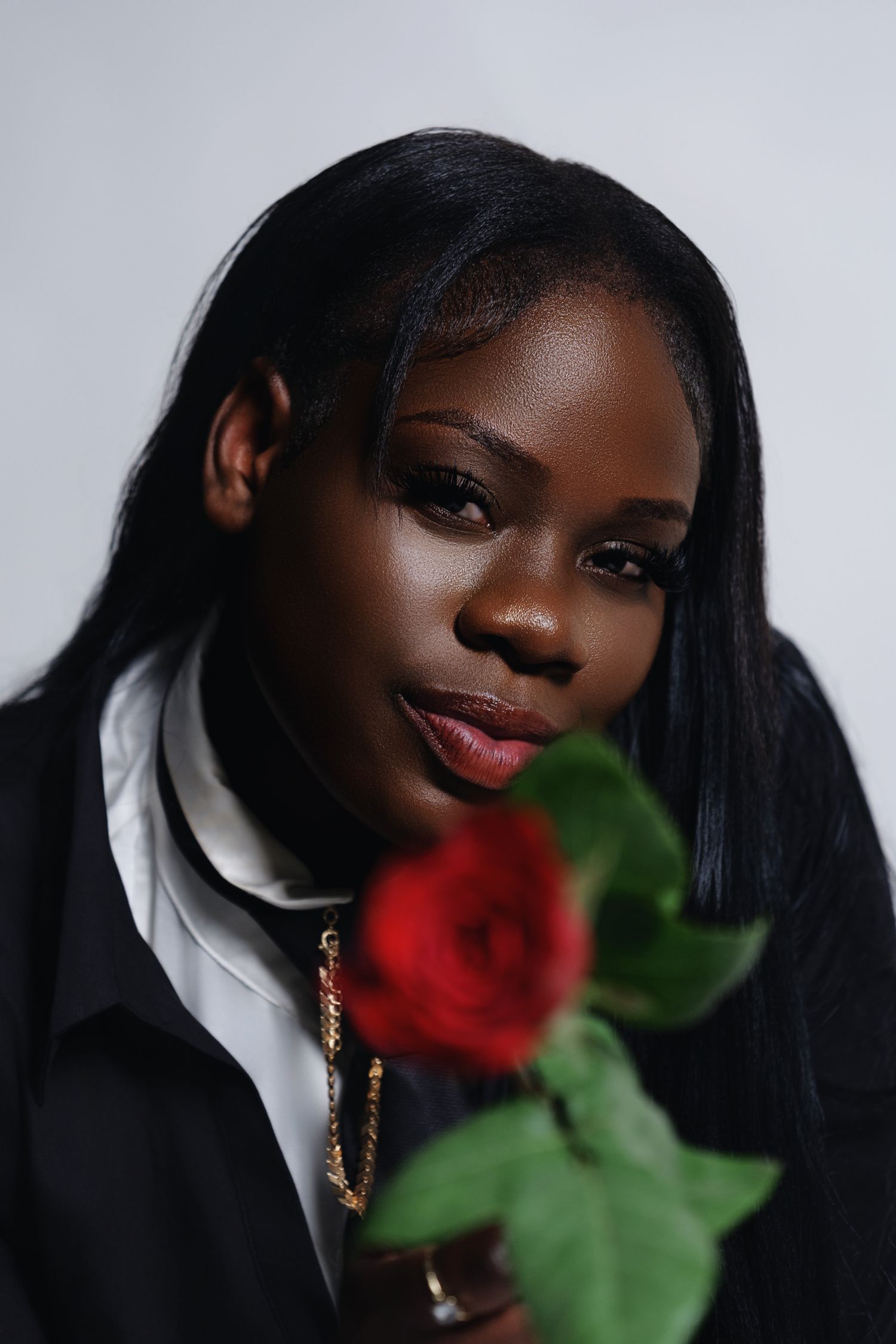 Young Black woman with straight hair holds a red rose, wearing a black jacket and gold necklace against gray background.