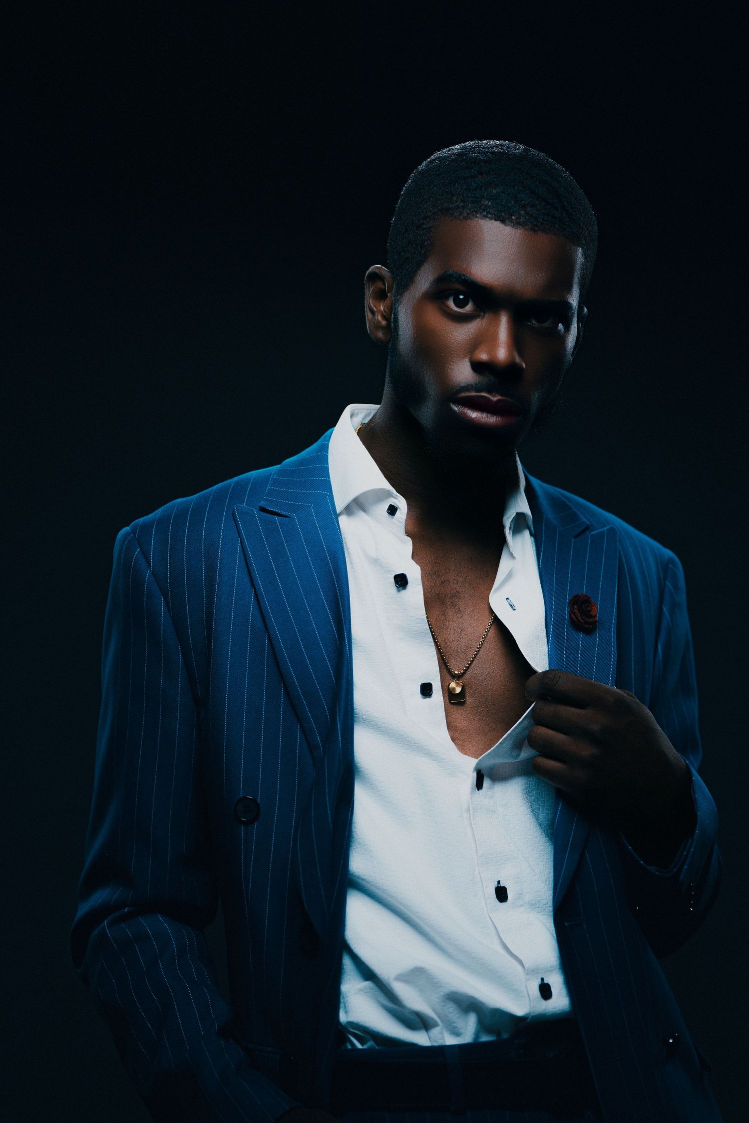 Stylish Black man in blue pinstripe suit and open white shirt with gold necklace poses confidently against dark background.