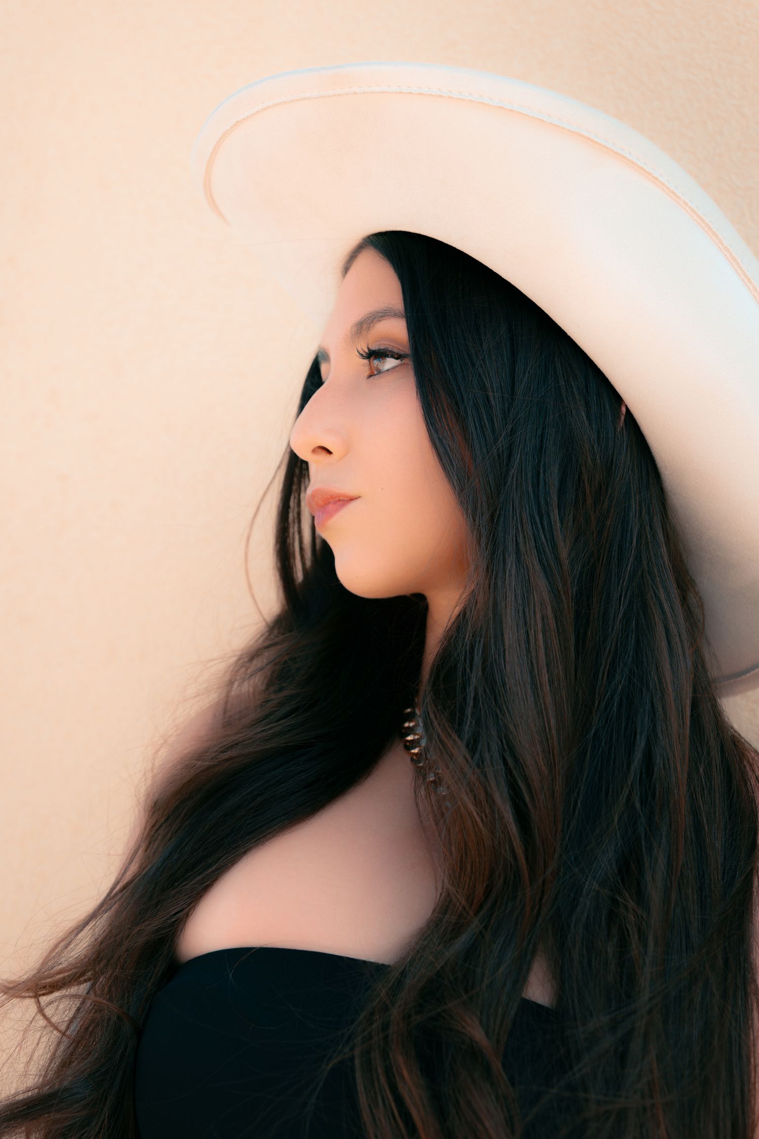 Young woman with long dark hair wearing a wide-brim white hat and black top, posing against a beige wall.