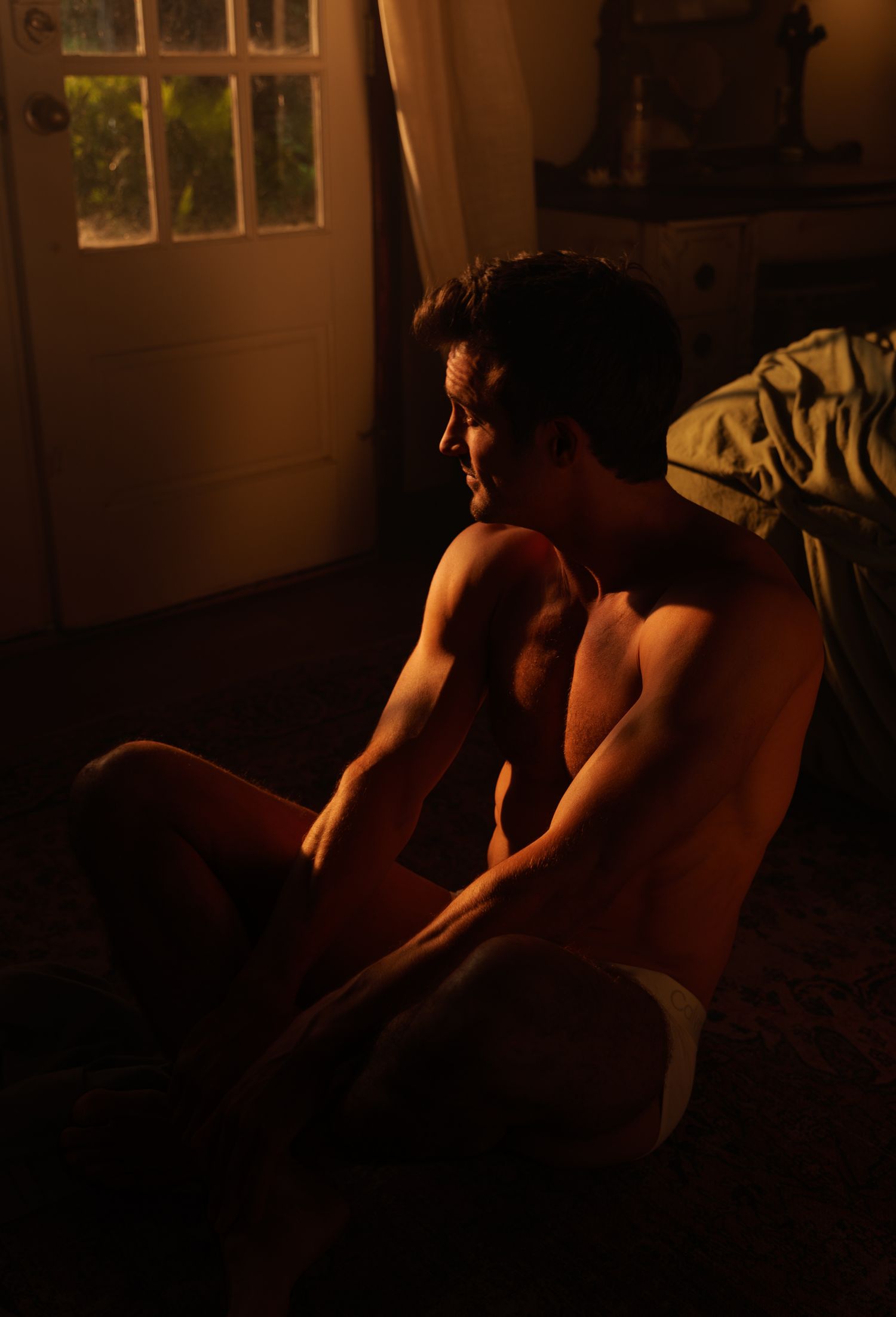 Shirtless muscular man sitting on floor in warm golden light near a window in a dimly lit room.