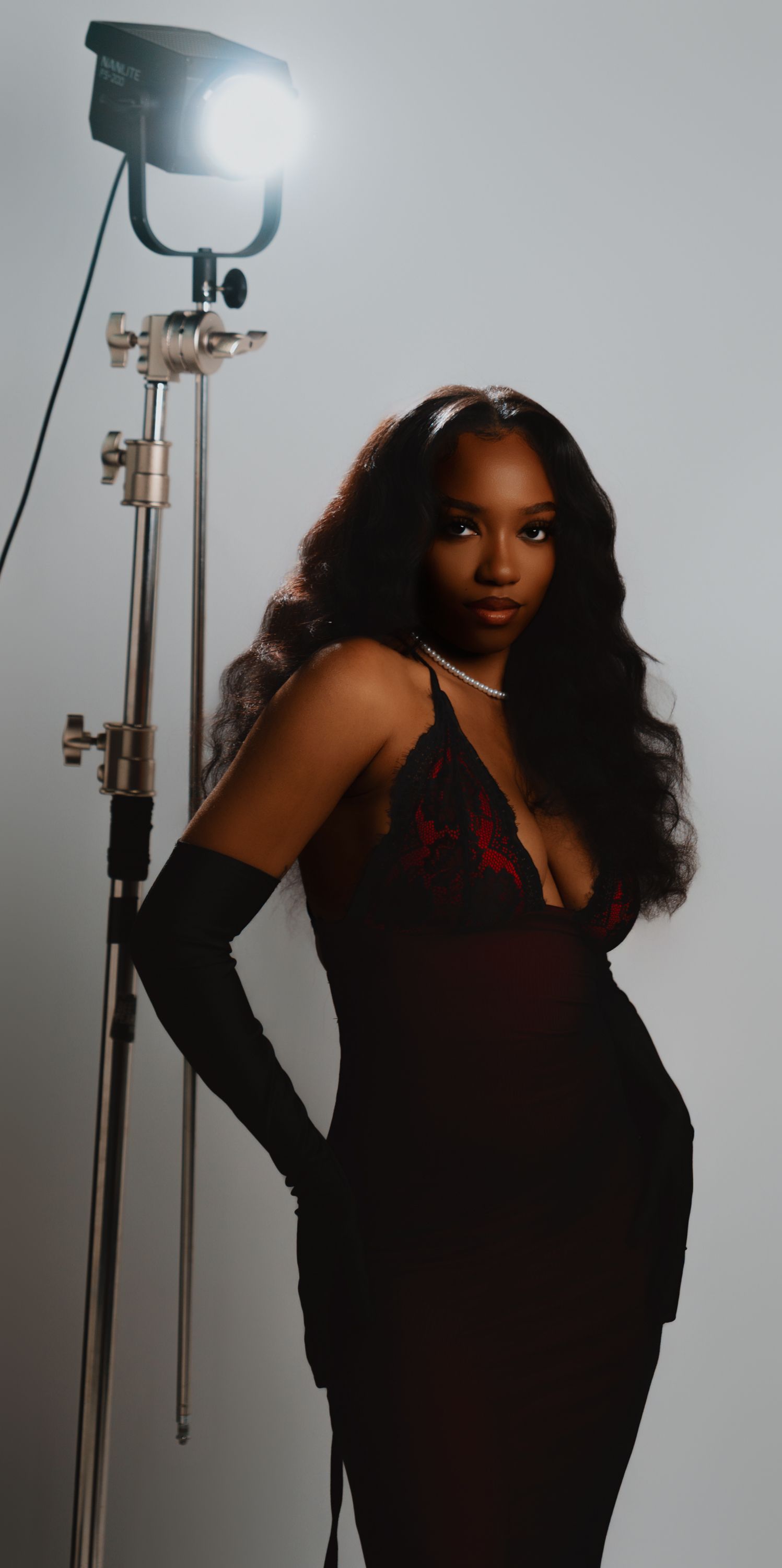 A woman in a black lace dress and long gloves poses elegantly beside a studio light against a white background.