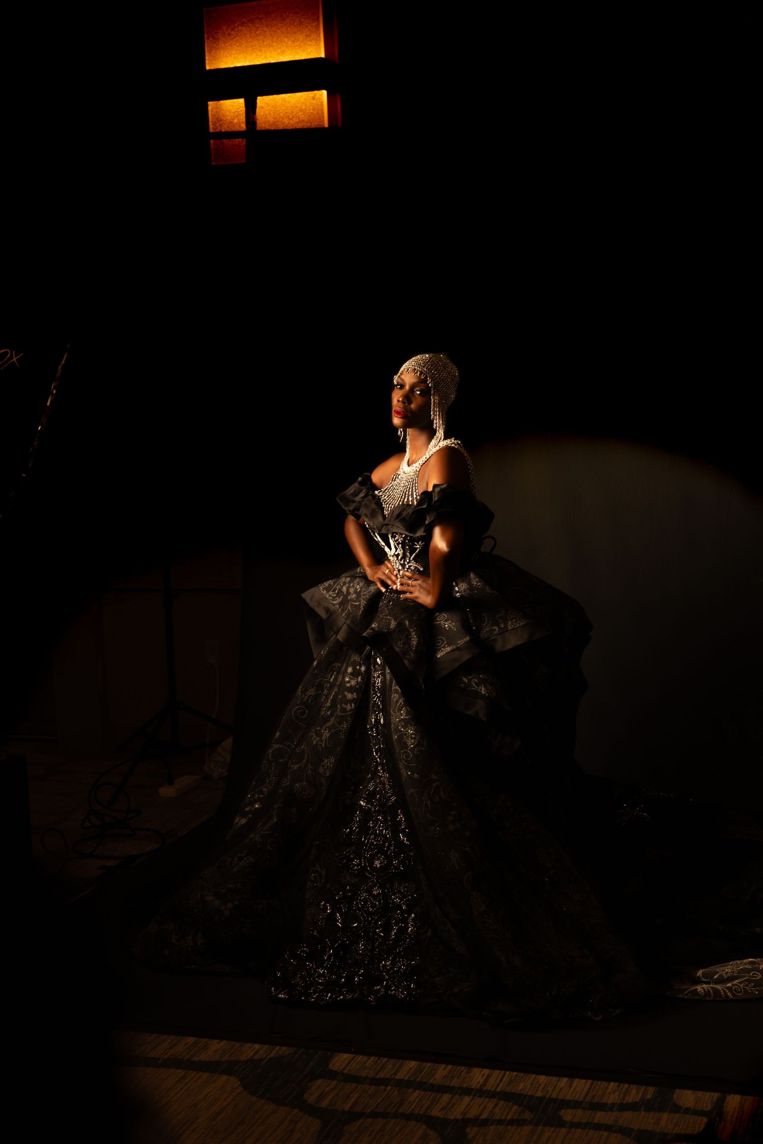 A woman in an elaborate dark gown and crown stands in dramatic low lighting, exuding regal elegance.