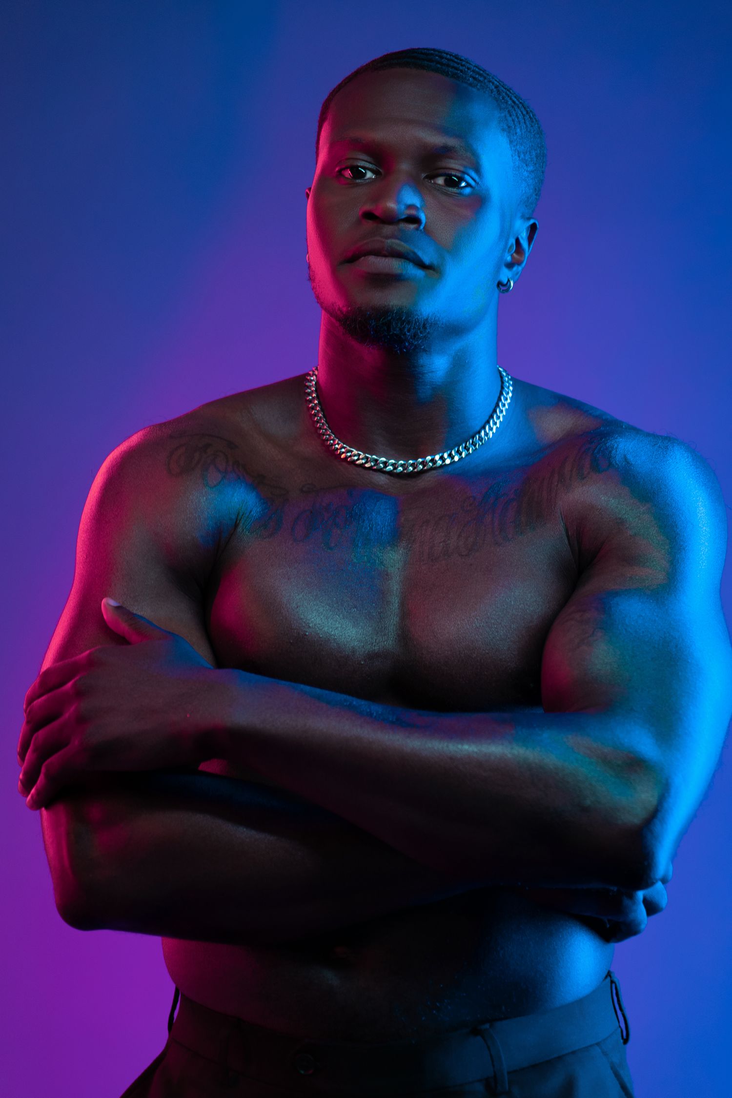 Muscular shirtless Black man with crossed arms, silver chain necklace, chest tattoos, posed against blue-purple neon background.