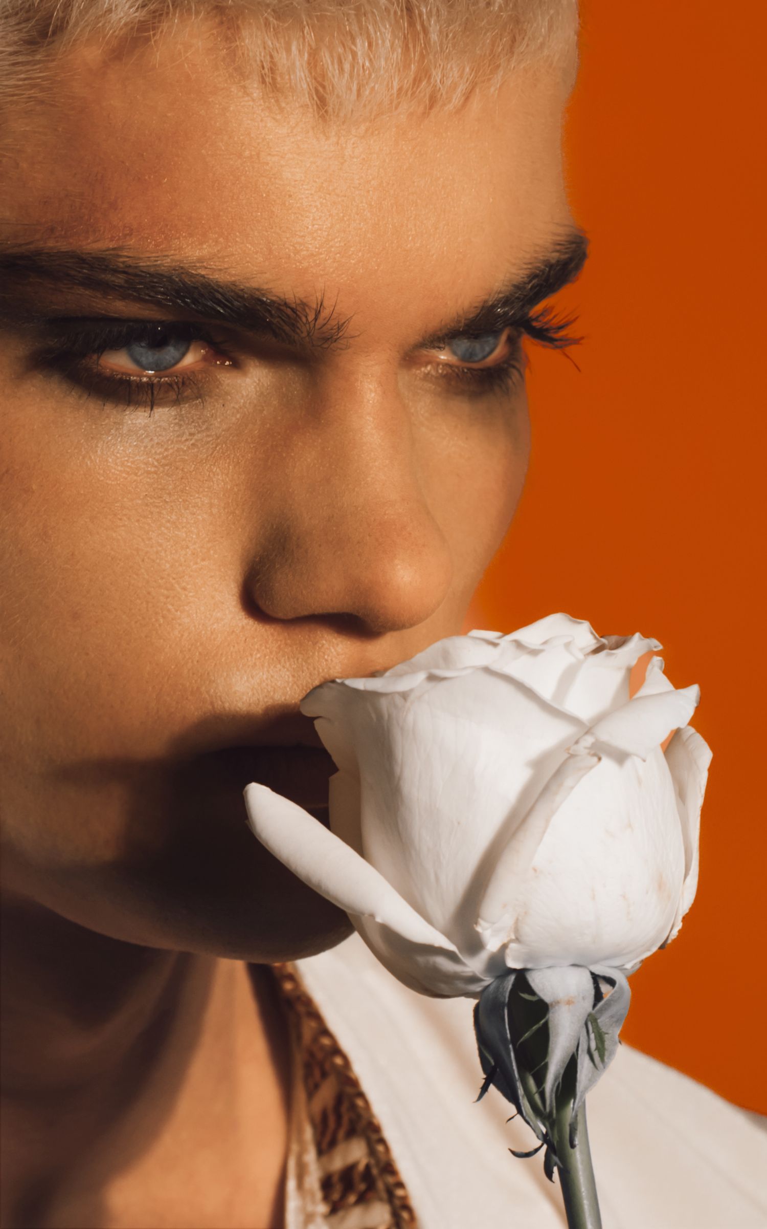 Young person with dramatic eye makeup smelling a white rose against a vibrant orange background.