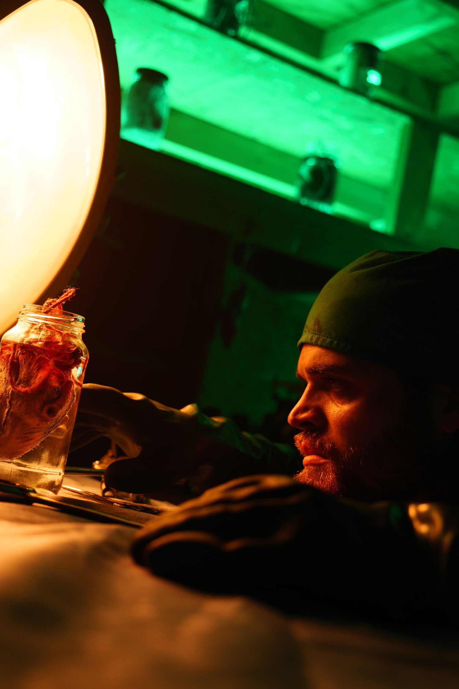 A man in a green cap examines a jar under warm lamp light in a dark, green-lit room.