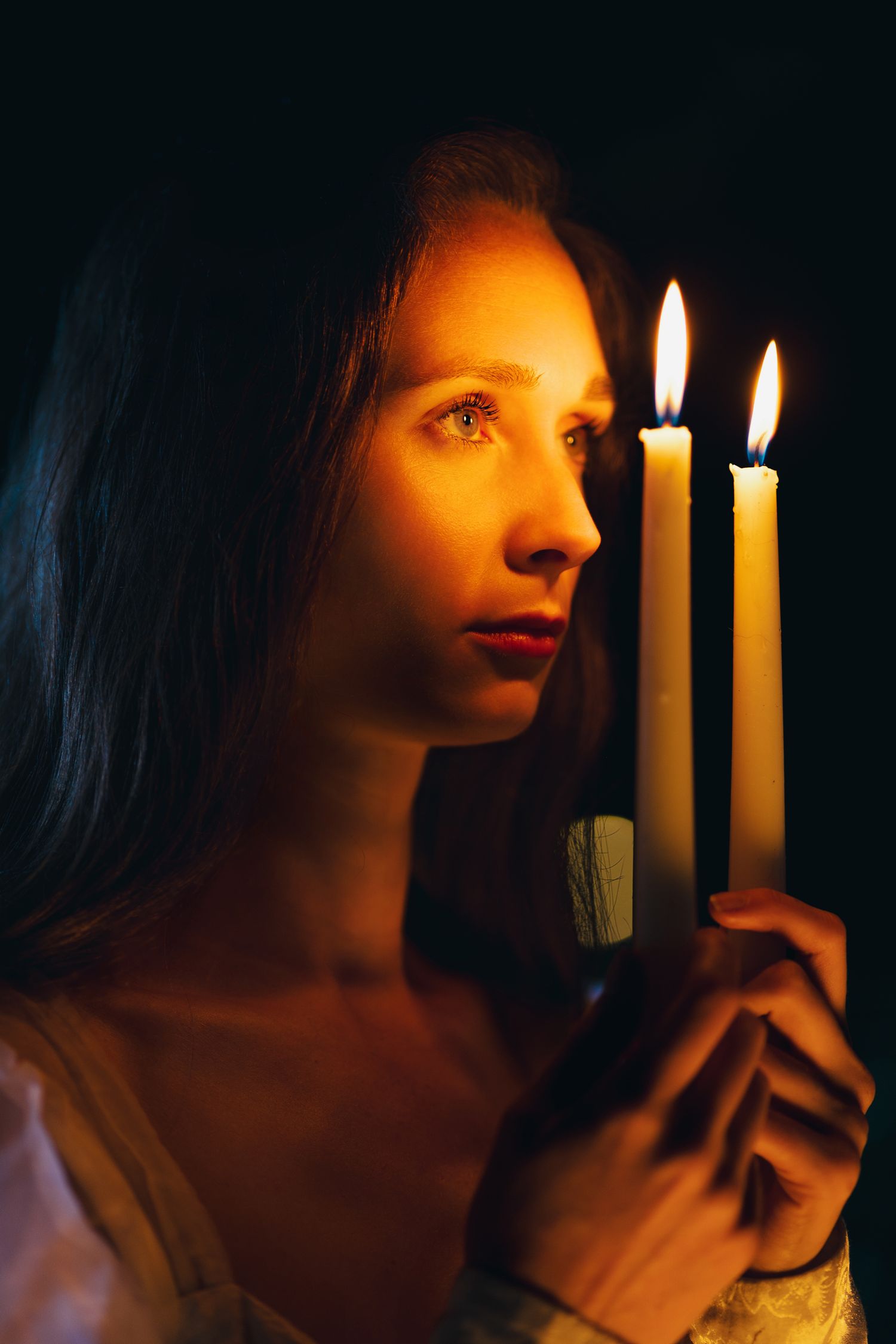 Young woman holding three lit candles in darkness, her face illuminated by warm golden candlelight.
