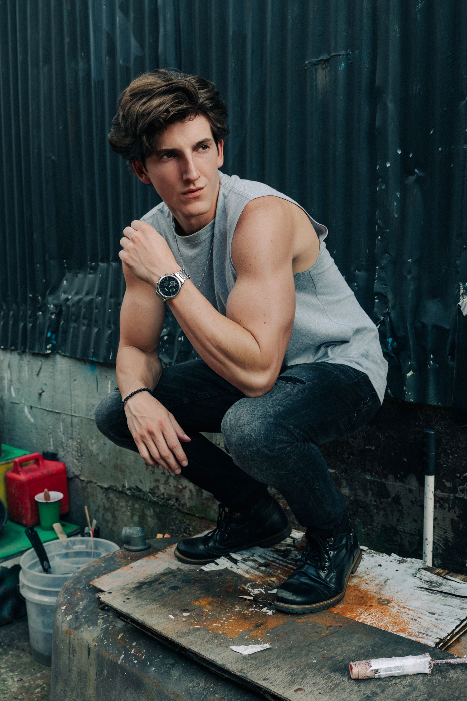 Young man in sleeveless shirt and black jeans crouching outdoors, wearing a watch, posed against a metal wall.