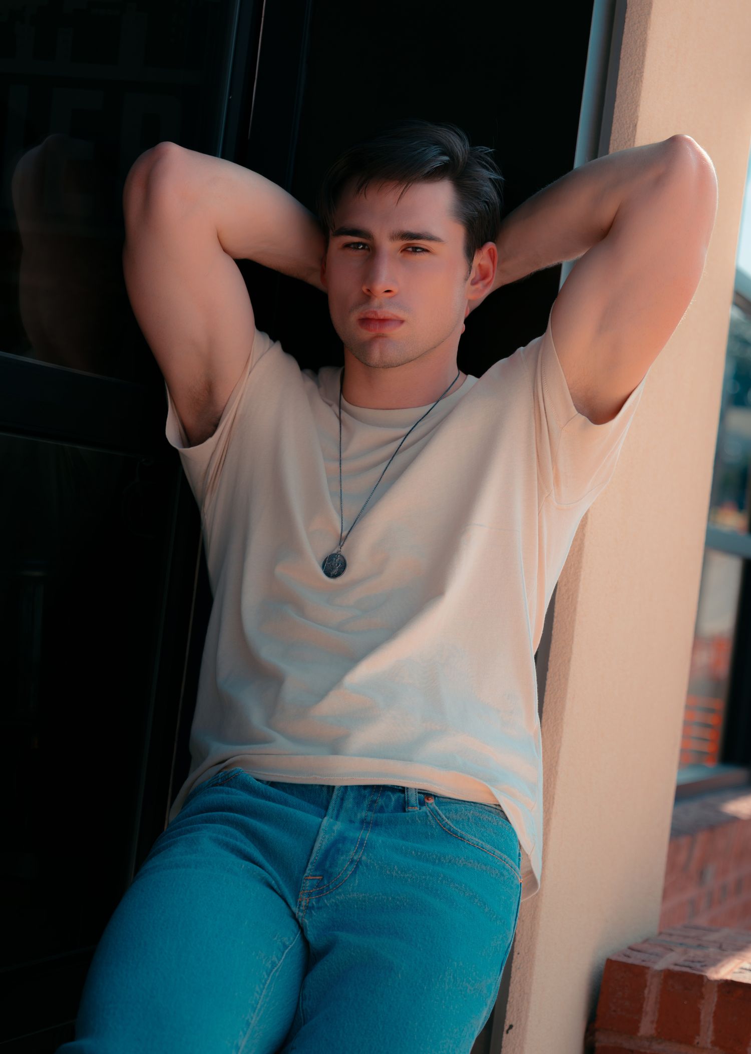 Young man in white t-shirt and jeans poses relaxed with hands behind head, wearing a pendant necklace outdoors.