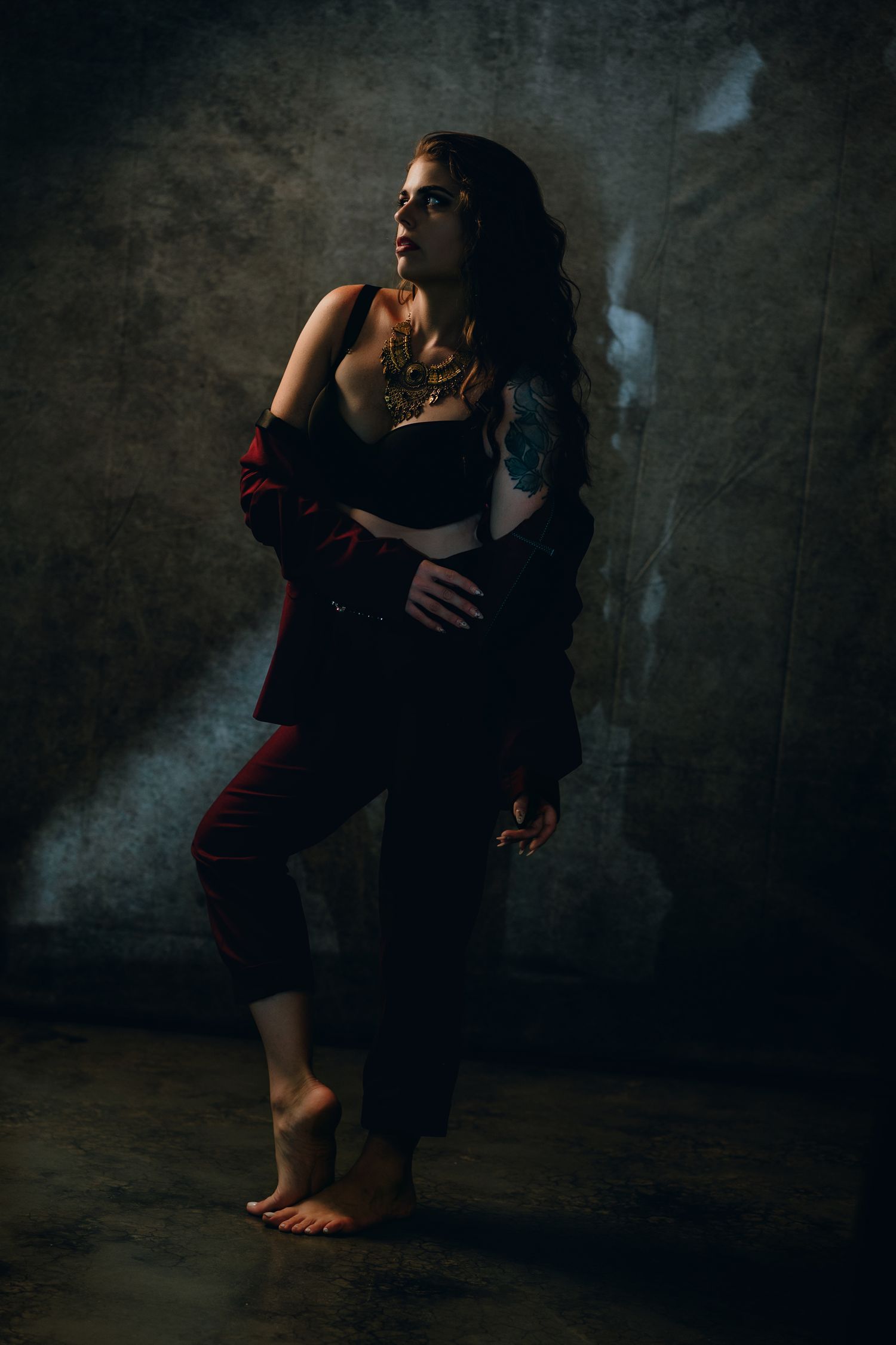 Tattooed woman in dark suit jacket and crop top poses barefoot against moody concrete wall in dramatic lighting.