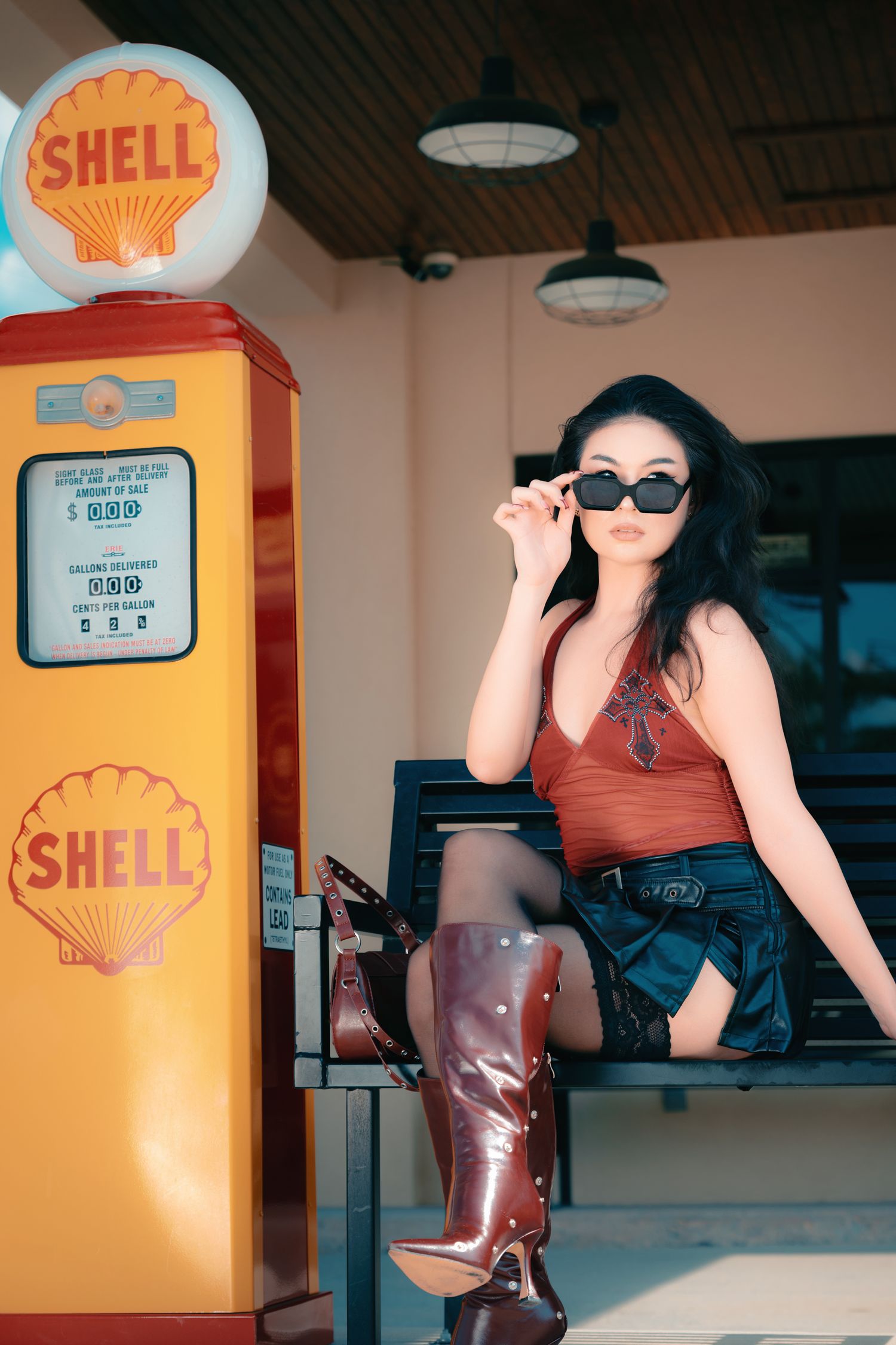 Woman in sunglasses and brown boots poses beside a vintage Shell gas pump at a retro gas station.
