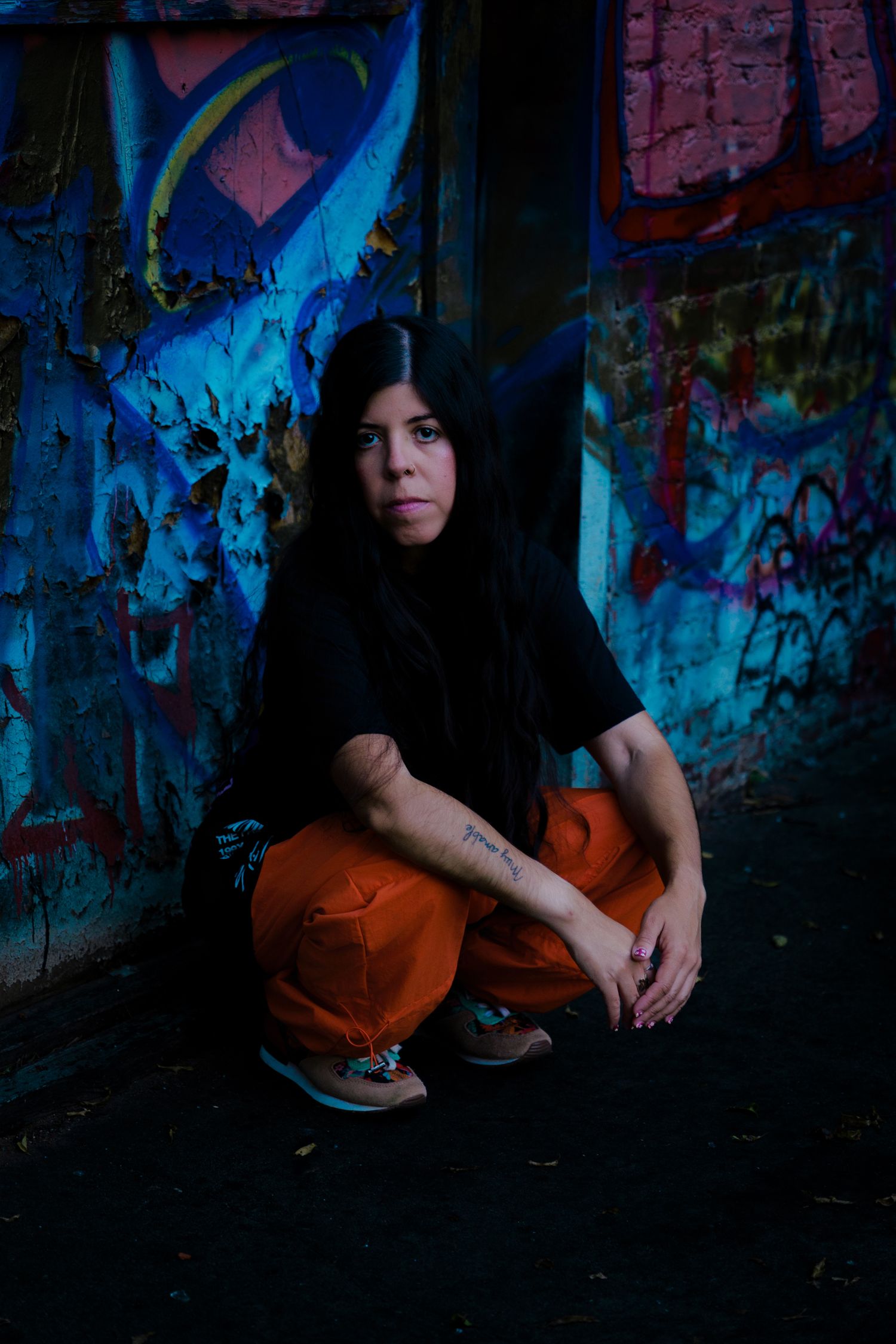 Woman in orange pants crouching against graffiti wall in urban setting.
