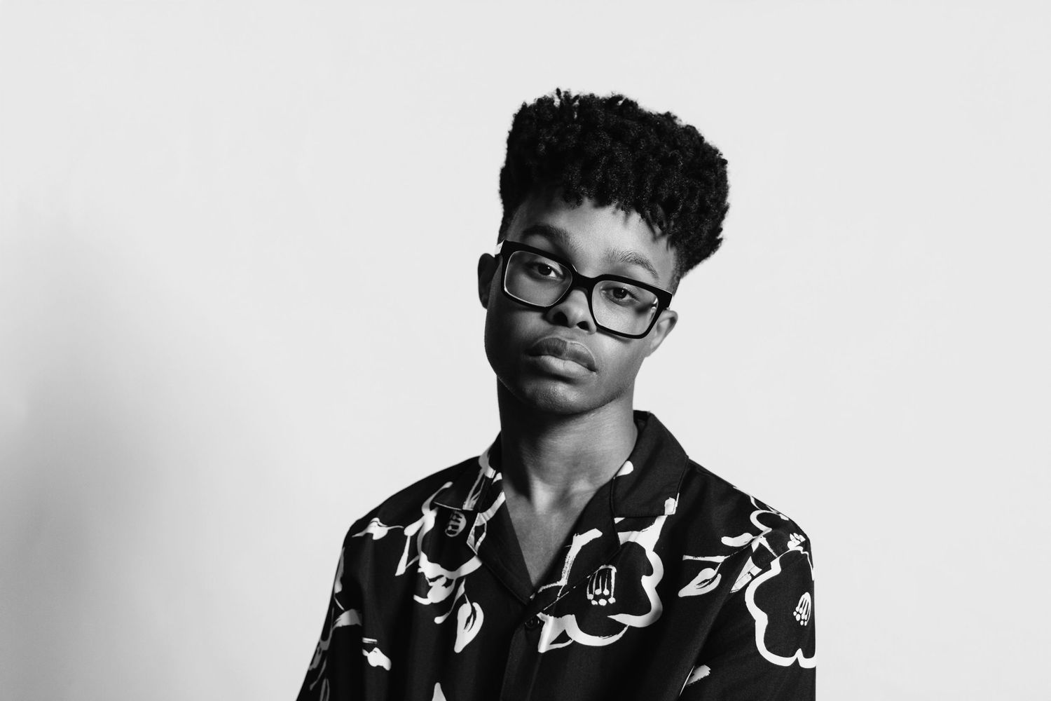 Black and white portrait of a young person with curly hair and glasses wearing a patterned shirt.