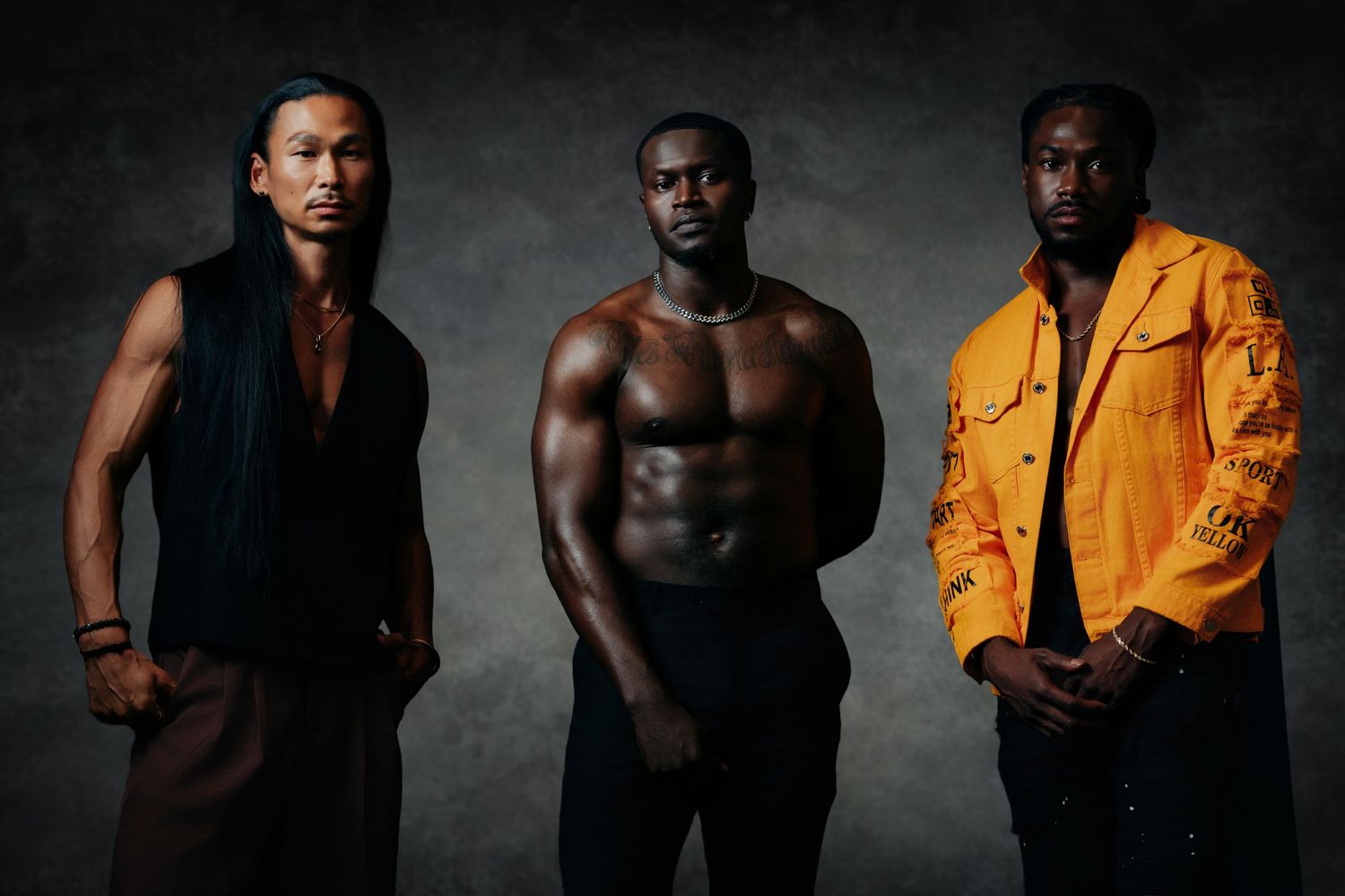 Three athletic men pose confidently in a studio, one shirtless showing muscular physique, flanked by two in stylish outfits.