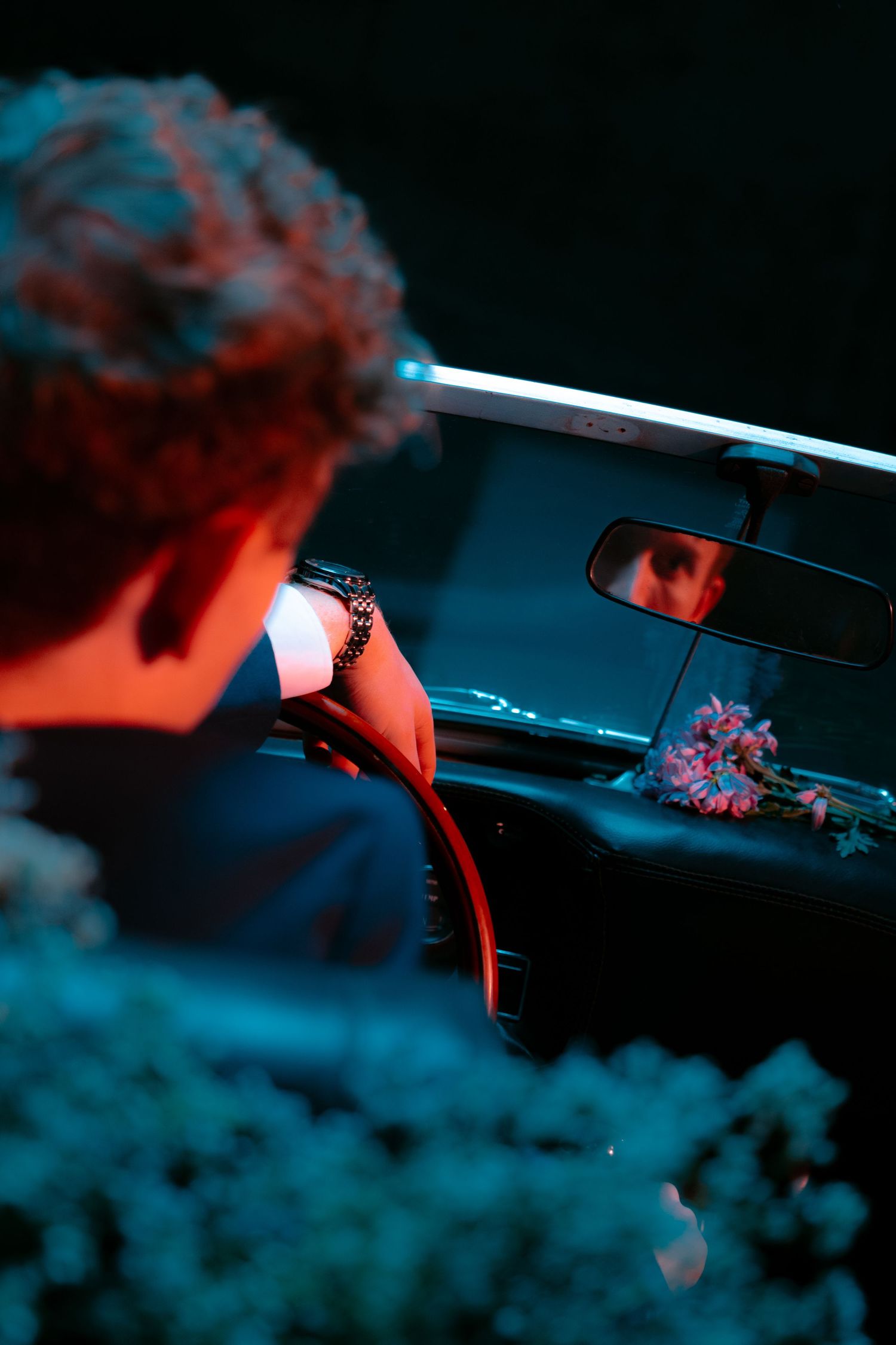Man with bracelet sits in dark car cockpit, flowers on dashboard, reflection visible in rearview mirror, red and blue lighting.