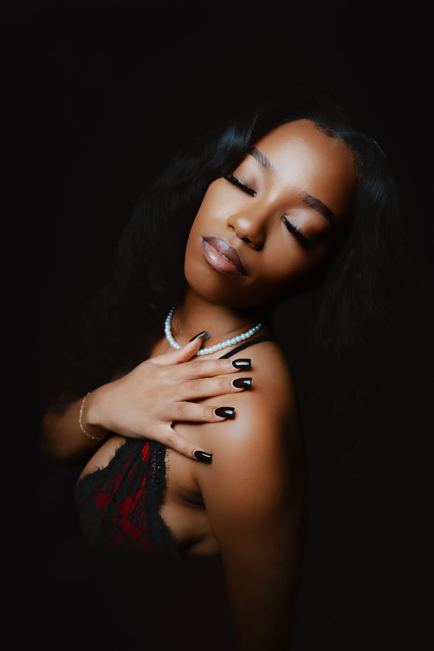 A Black woman with long hair, black nails, and pearl necklace poses gracefully against a dark background.