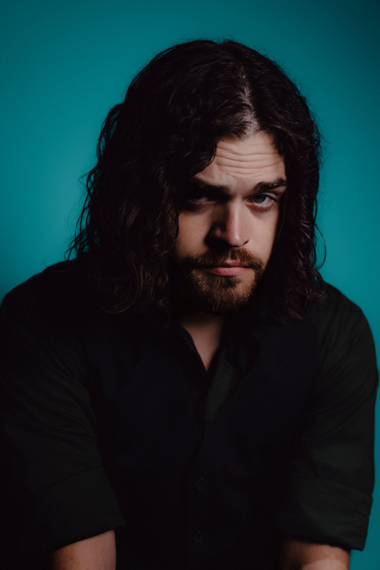 Young man with long dark curly hair and beard wearing black shirt poses against teal background.