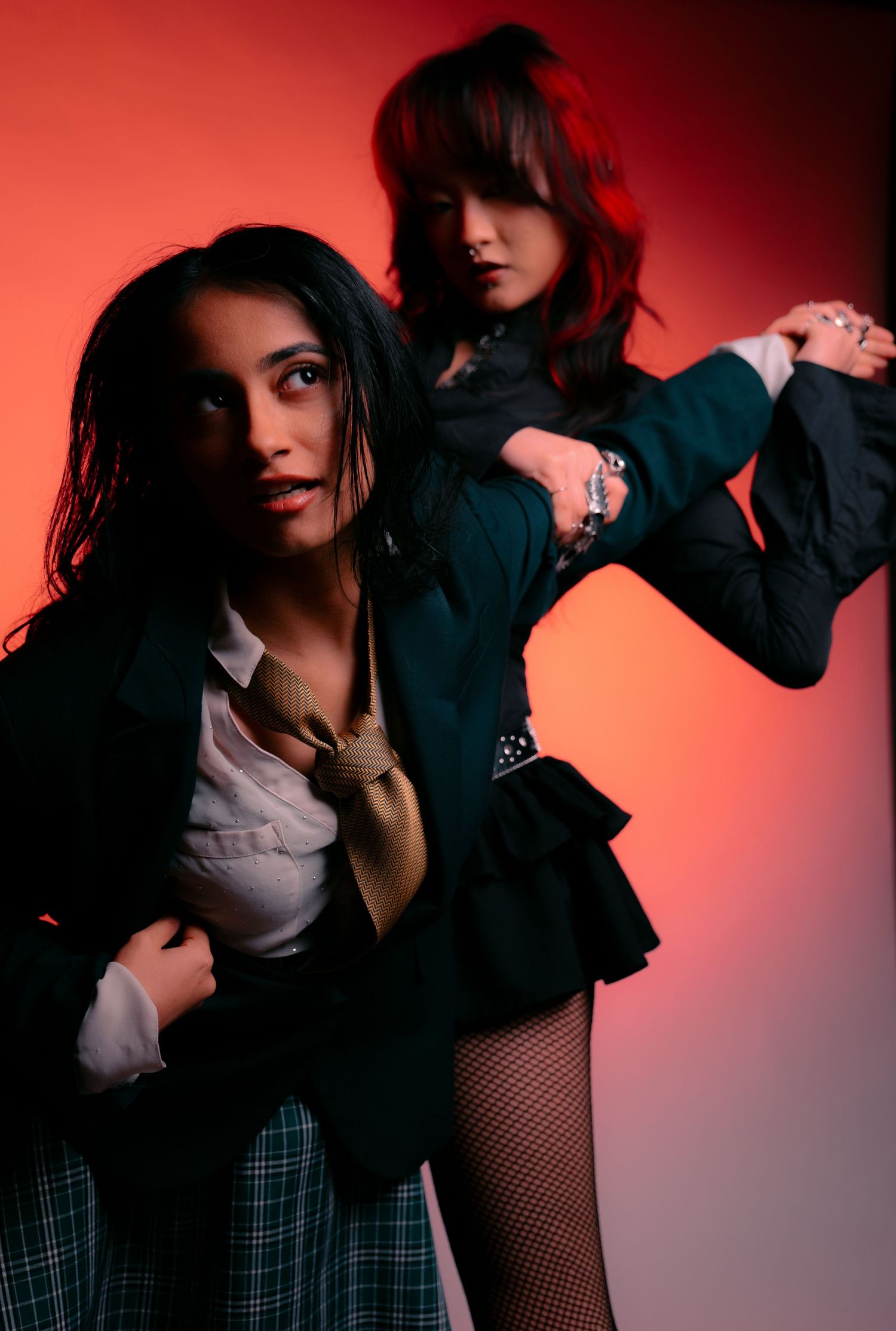 Two women in dark blazers and plaid skirts pose dramatically against a red background in edgy fashion shoot.