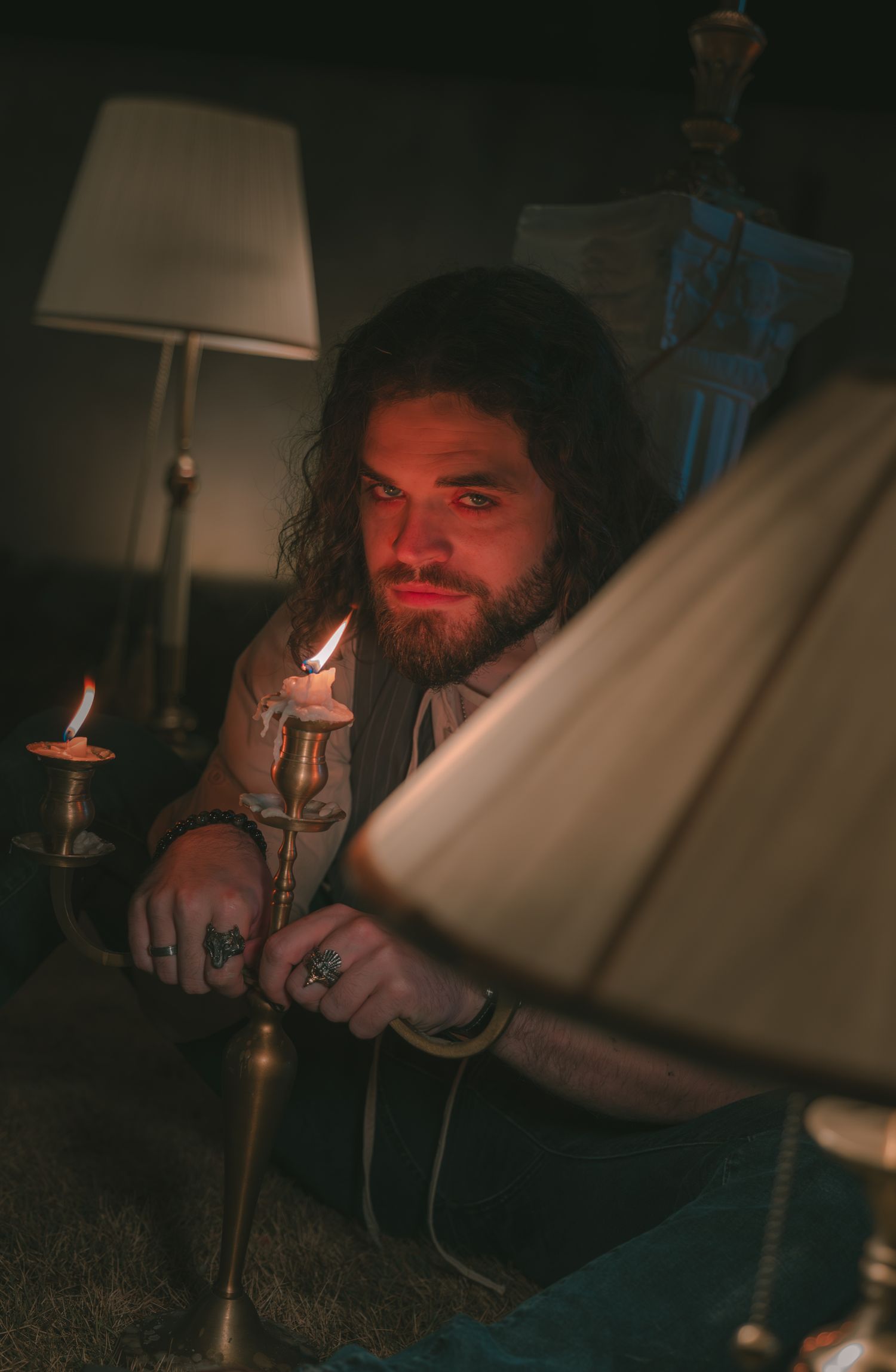 Bearded man with long hair holding a lit candle in a dimly lit room with tattoos on his hands.