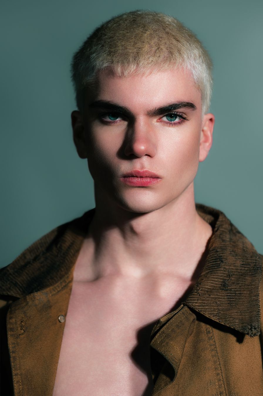 Young male model with platinum blonde hair, blue eyes, red lips, and brown jacket poses against teal background.