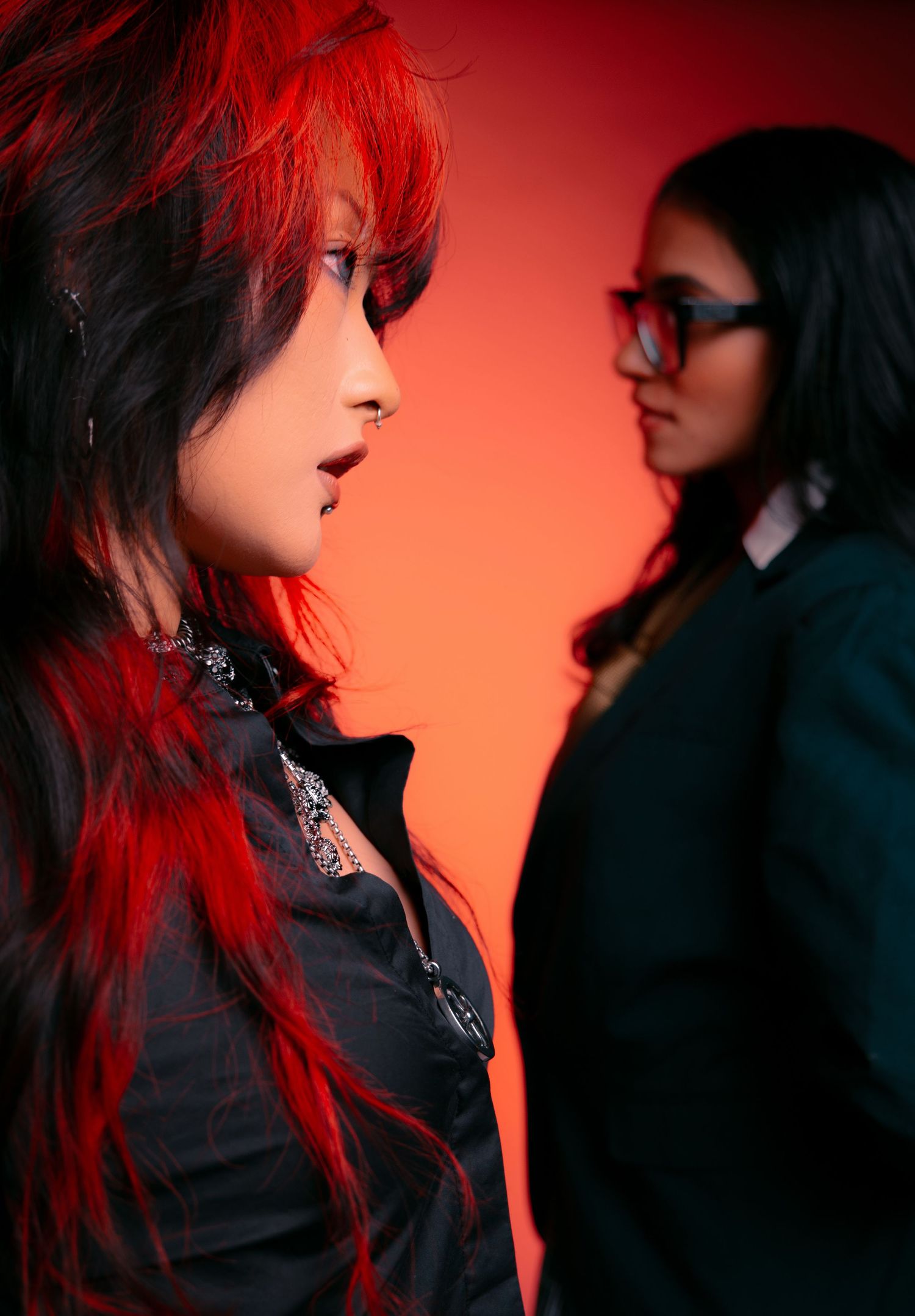 Two women with red-tinted hair pose dramatically against a red background, one in foreground with vibrant red-black hair.