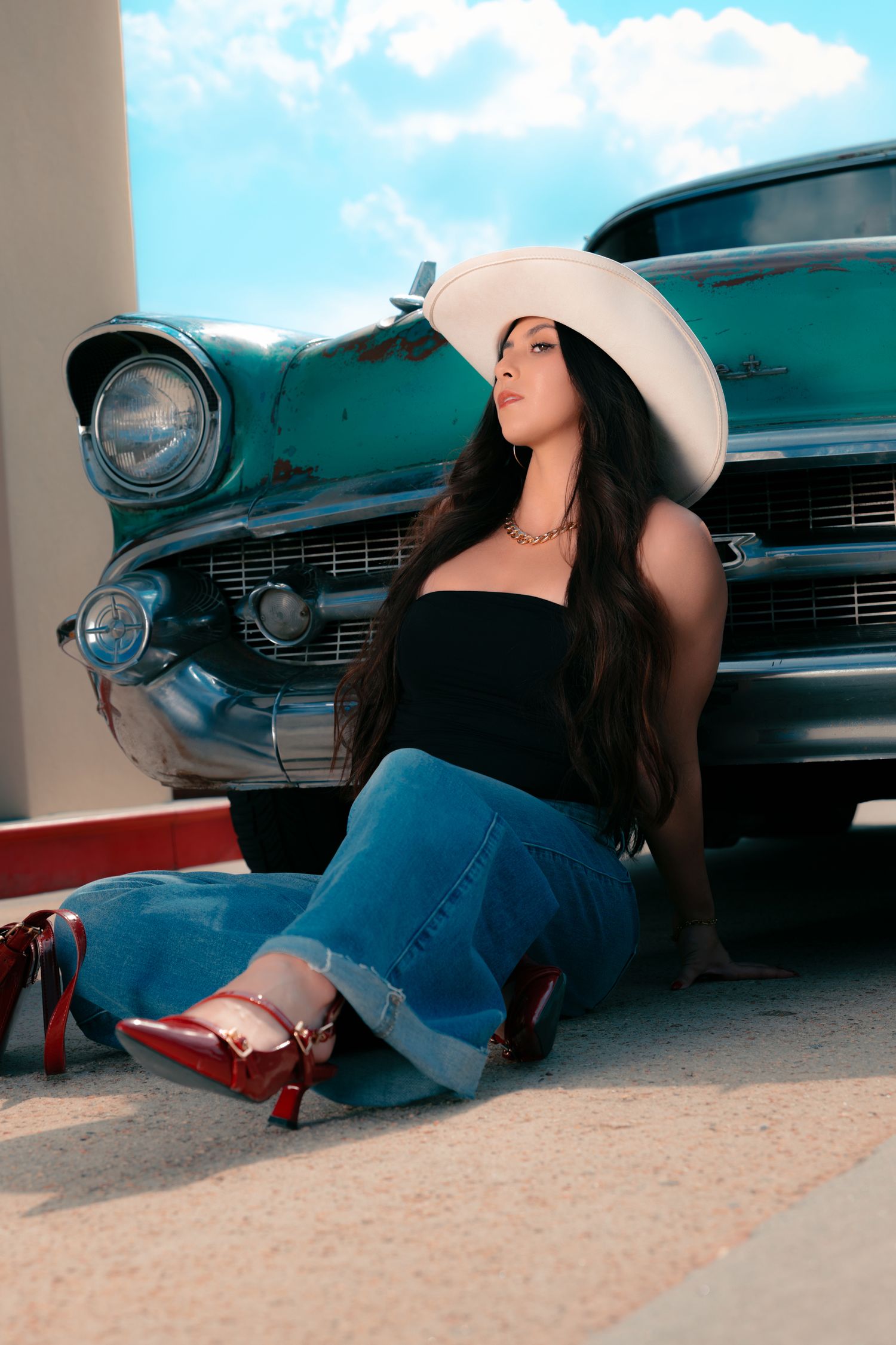 Woman in white cowboy hat, black top, jeans, and red heels sits beside a vintage teal classic car under blue sky.