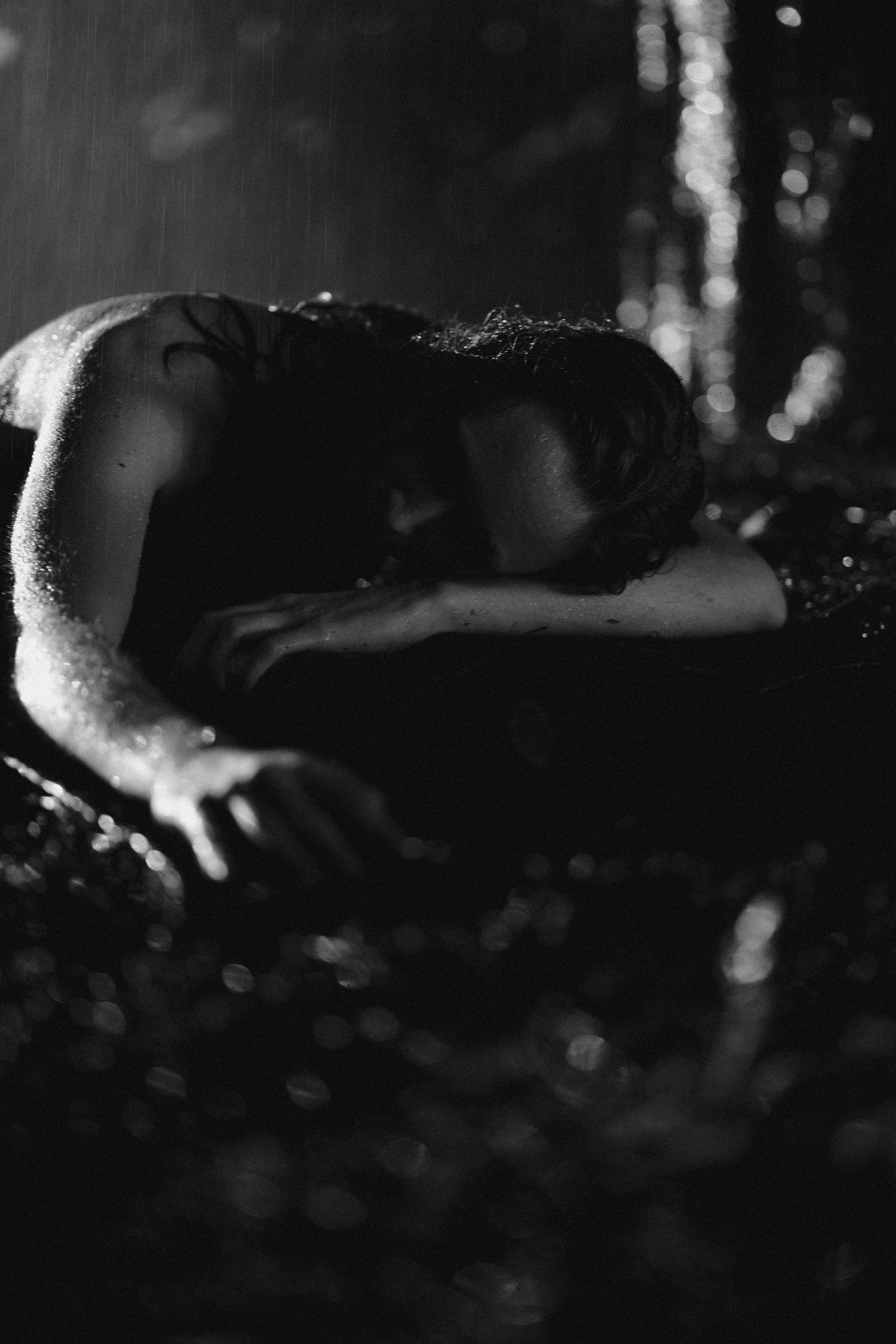 Black and white photo of a person resting their head on their arms in the rain, evoking emotion and solitude.