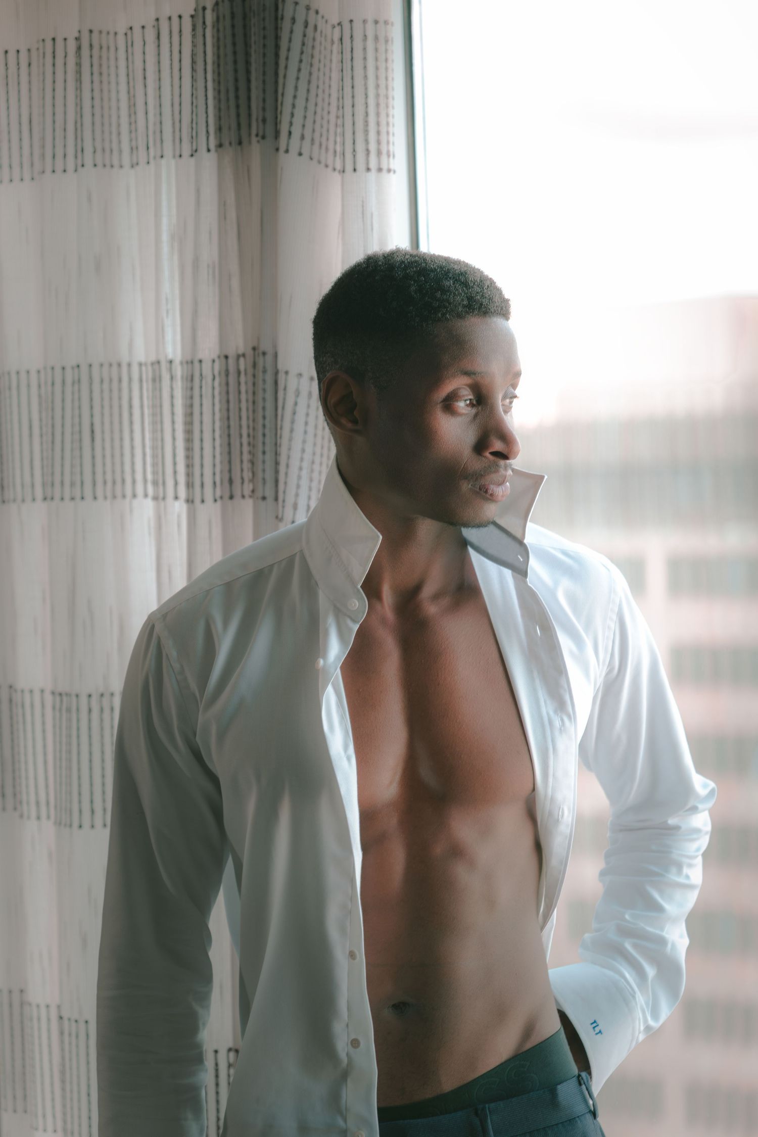 A fit young Black man stands by a window in an open white dress shirt, gazing to the side in soft natural light.