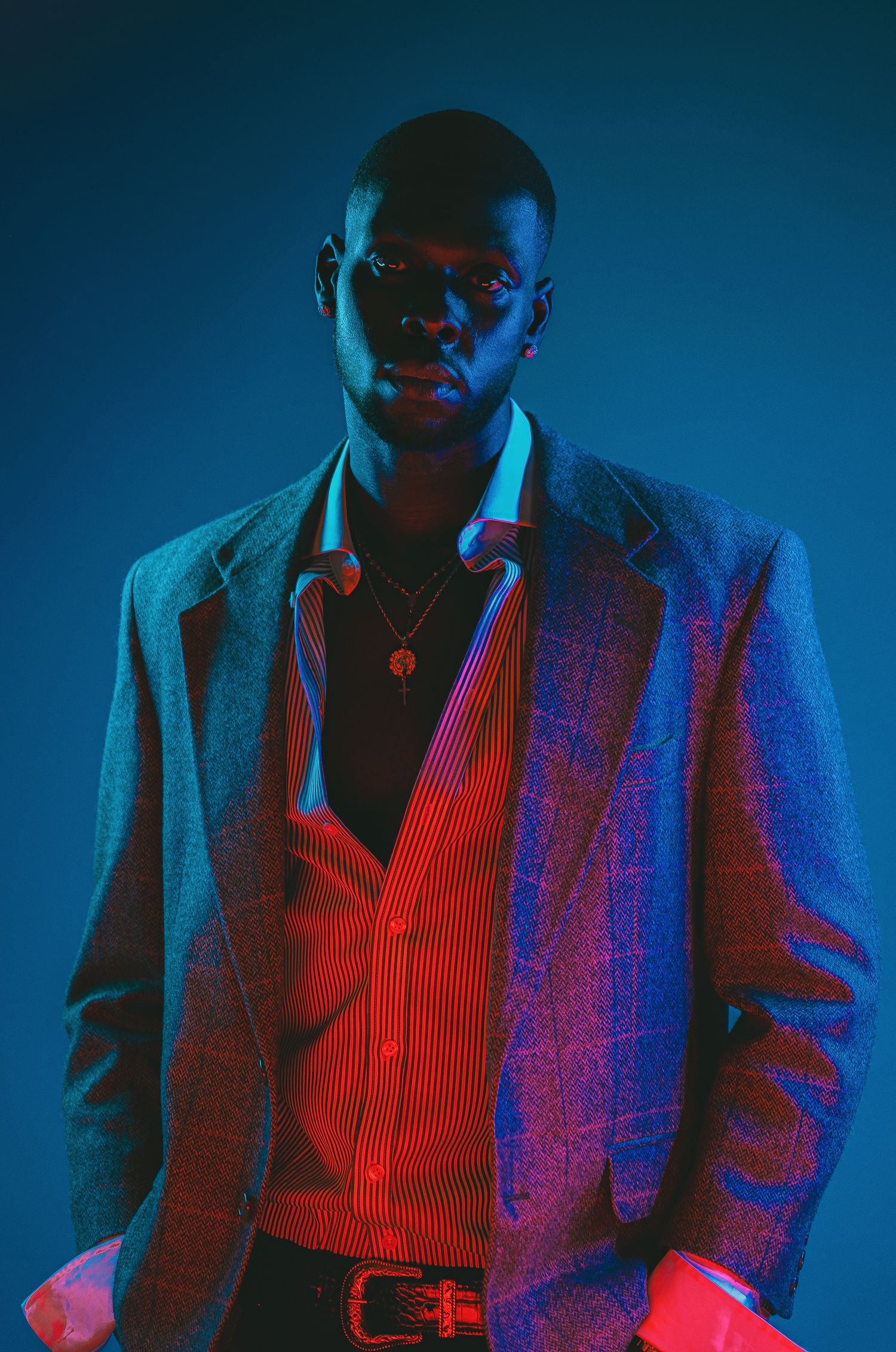 Young man in red striped shirt and gray blazer poses confidently under dramatic blue and red studio lighting.