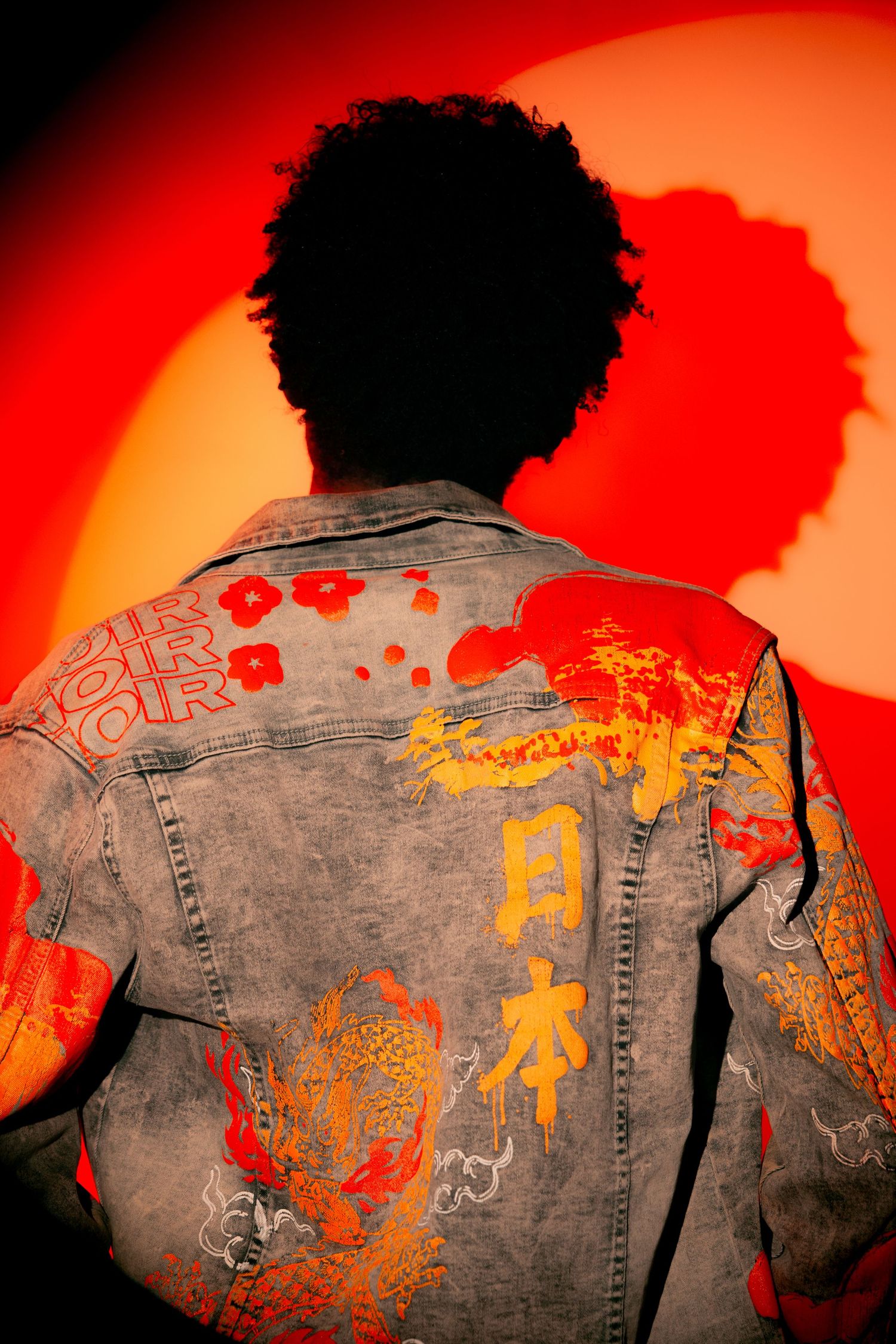 Person wearing painted denim jacket with Japanese kanji and dragon artwork, posed against red-orange background.
