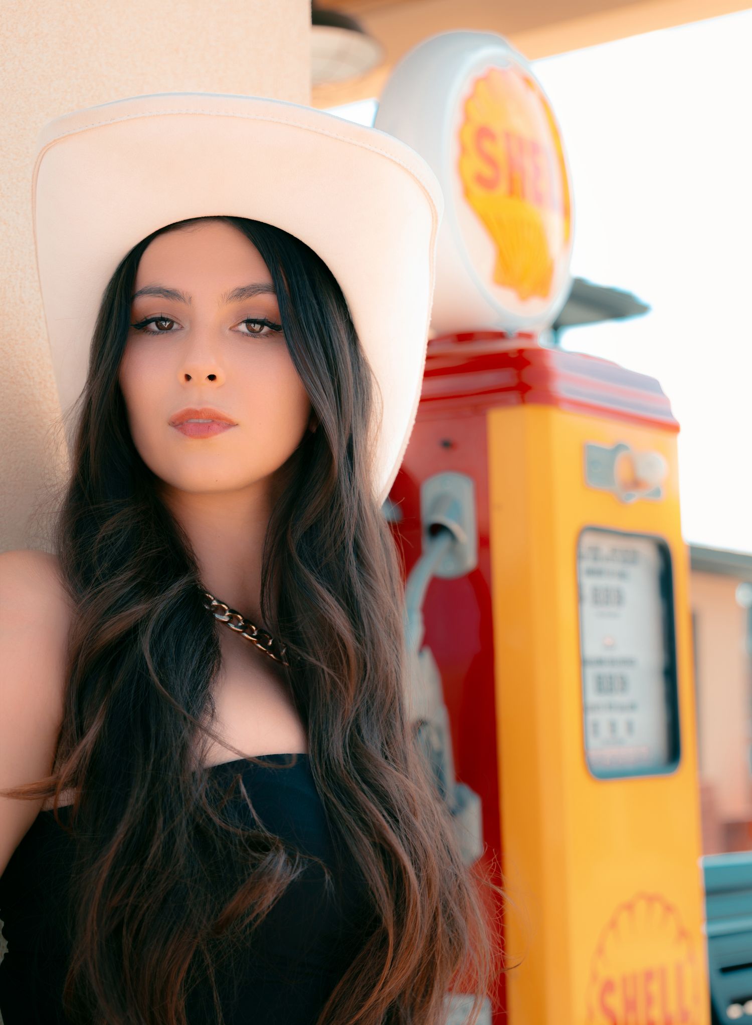 Woman in white cowboy hat with long dark hair poses near a vintage Shell gas pump in a retro outdoor setting.
