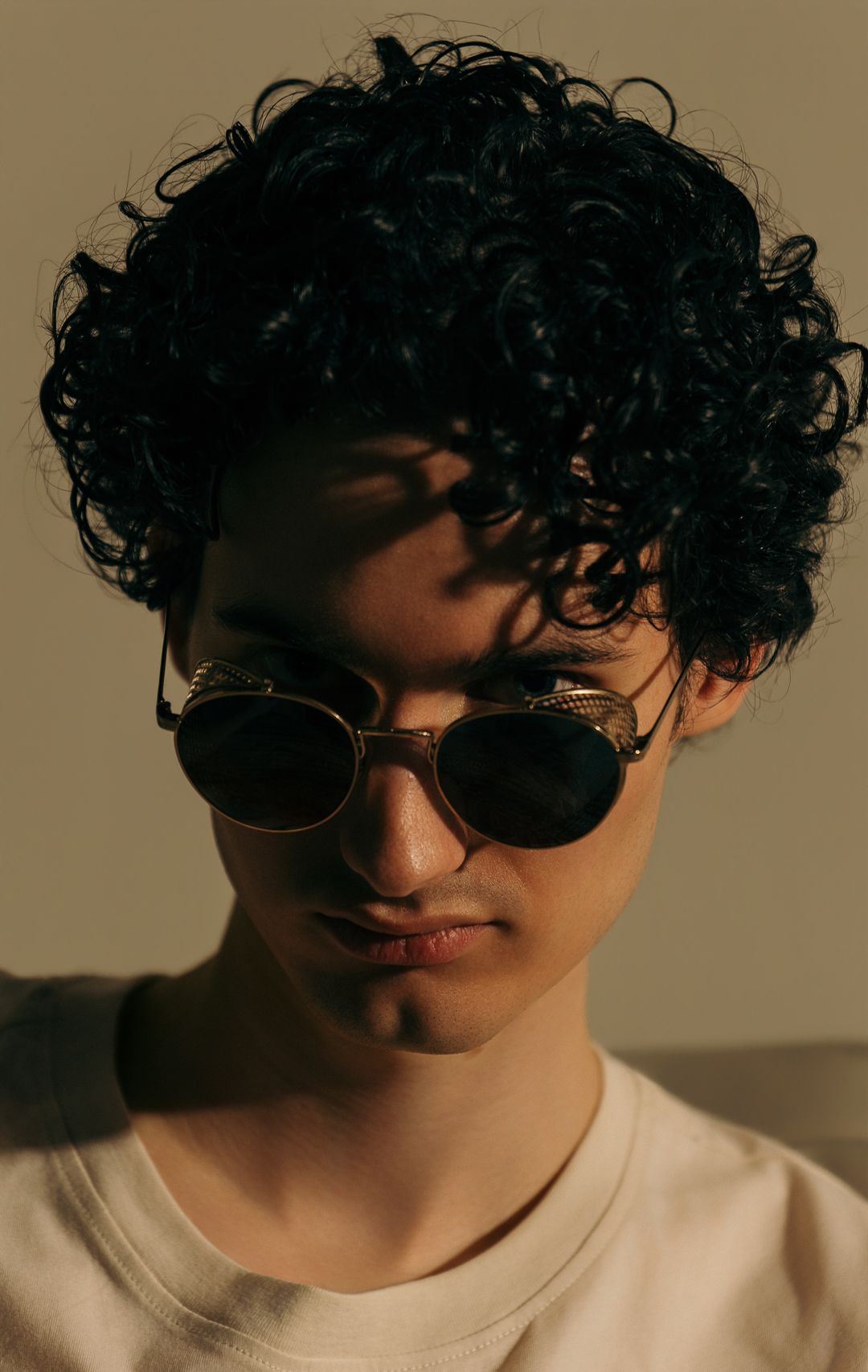 Young man with curly dark hair wearing gold aviator sunglasses in dramatic natural lighting.